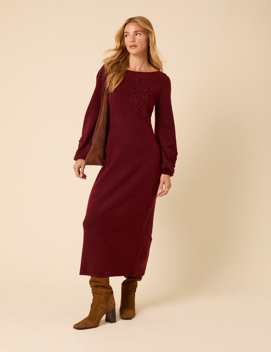 A woman wearing a burgundy long sleeve midi dress with floral embroidery, round neckline, and fitted style, paired with brown boots.