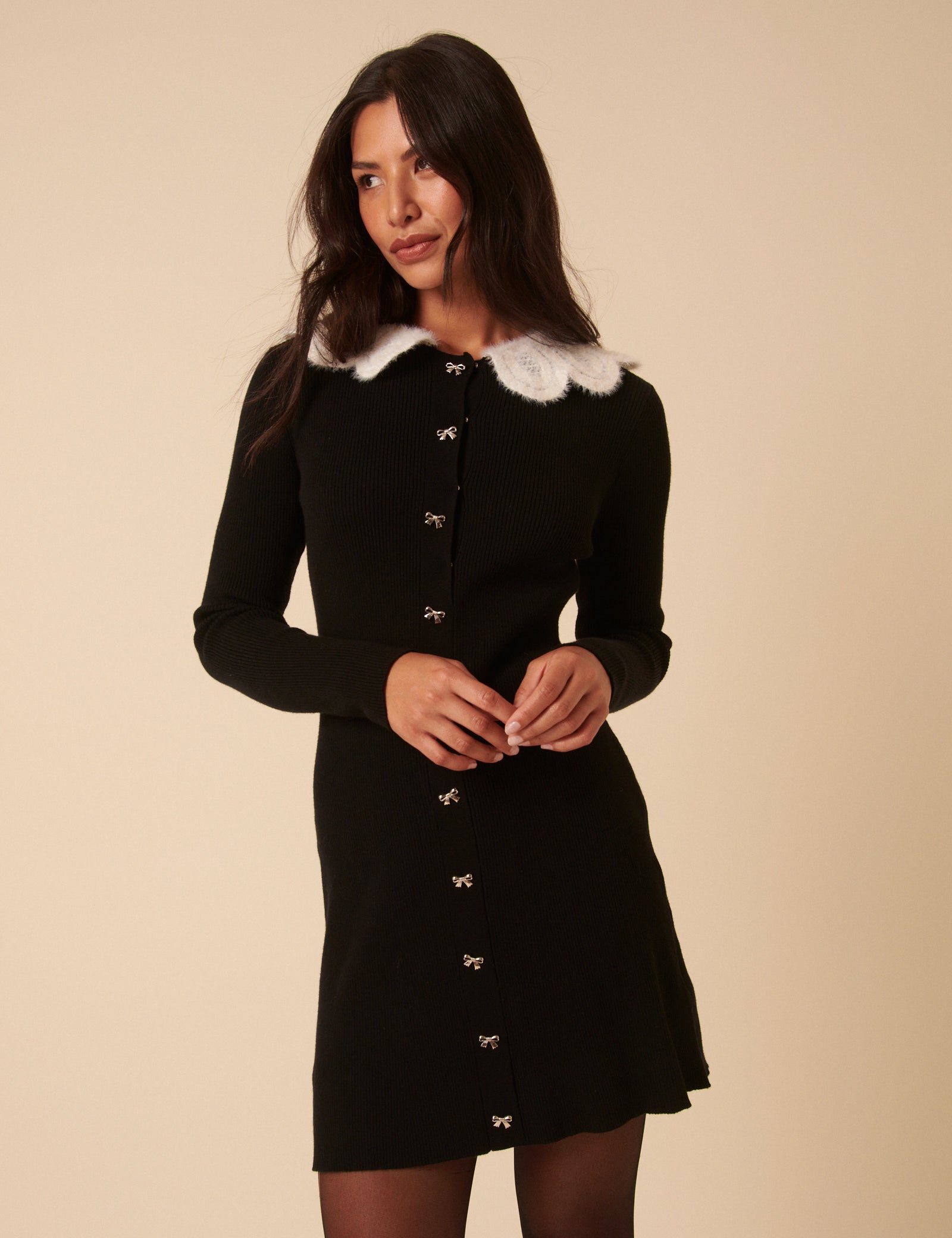 A woman wearing a black mini dress with a white crochet collar, long sleeves, and silver bow buttons down the front.