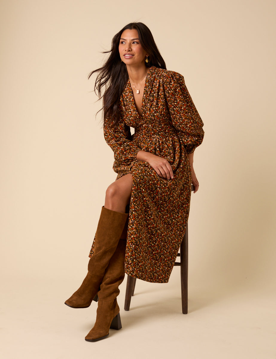 A woman wearing a black midi dress with a floral ditsy pattern, long sleeves, and a V-neck, sitting on a stool.