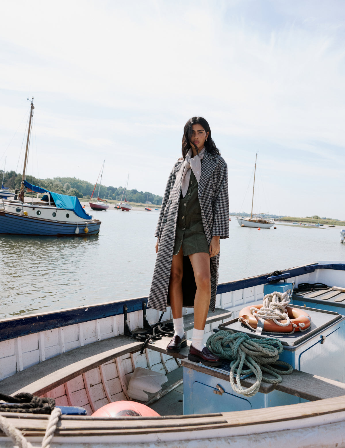 A woman wearing a black and brown houndstooth relaxed coat with a collar, long sleeves, flap pockets, and button fastening, standing on a boat with a marina in the background.