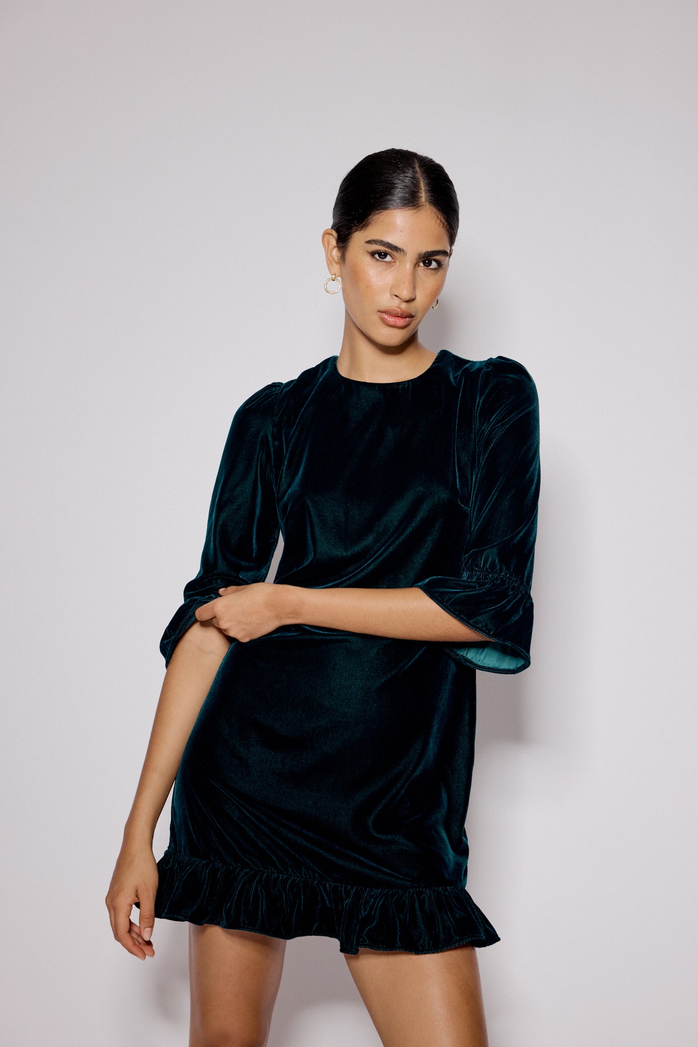 A woman wearing a dark green velvet mini dress with a round neckline, half-length sleeves, and ruffle trims at the cuffs and hem.
