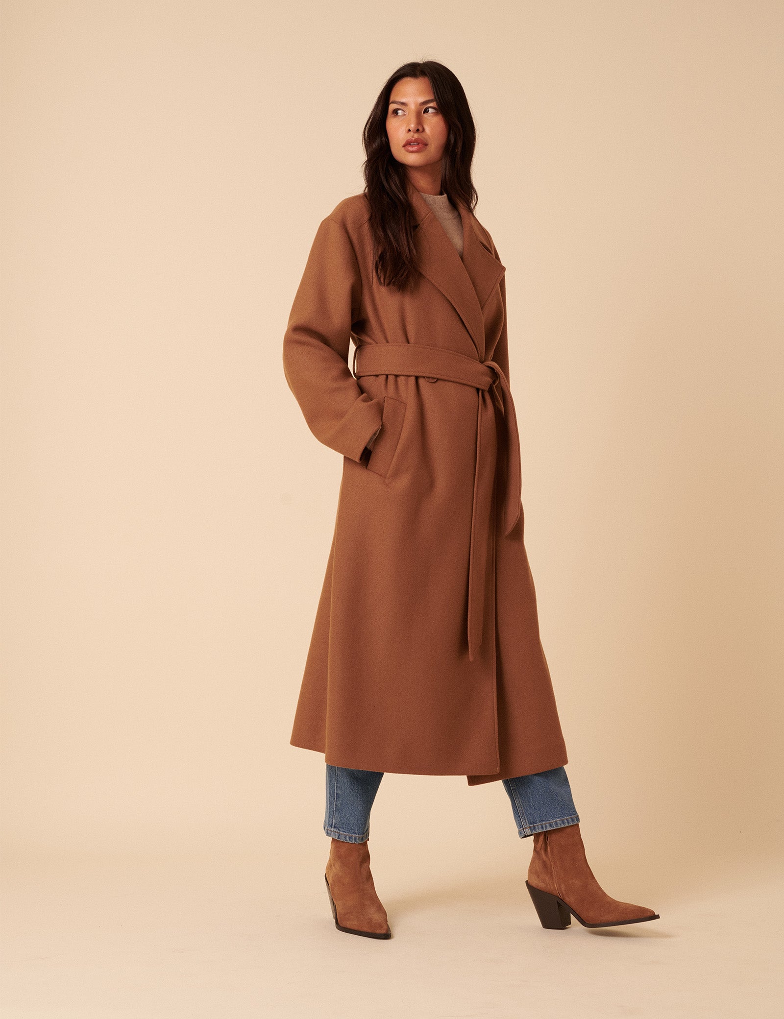 A woman wearing a camel brown wrap coat with a waist tie belt, classic collar, and side pockets, paired with blue jeans and brown heeled boots.