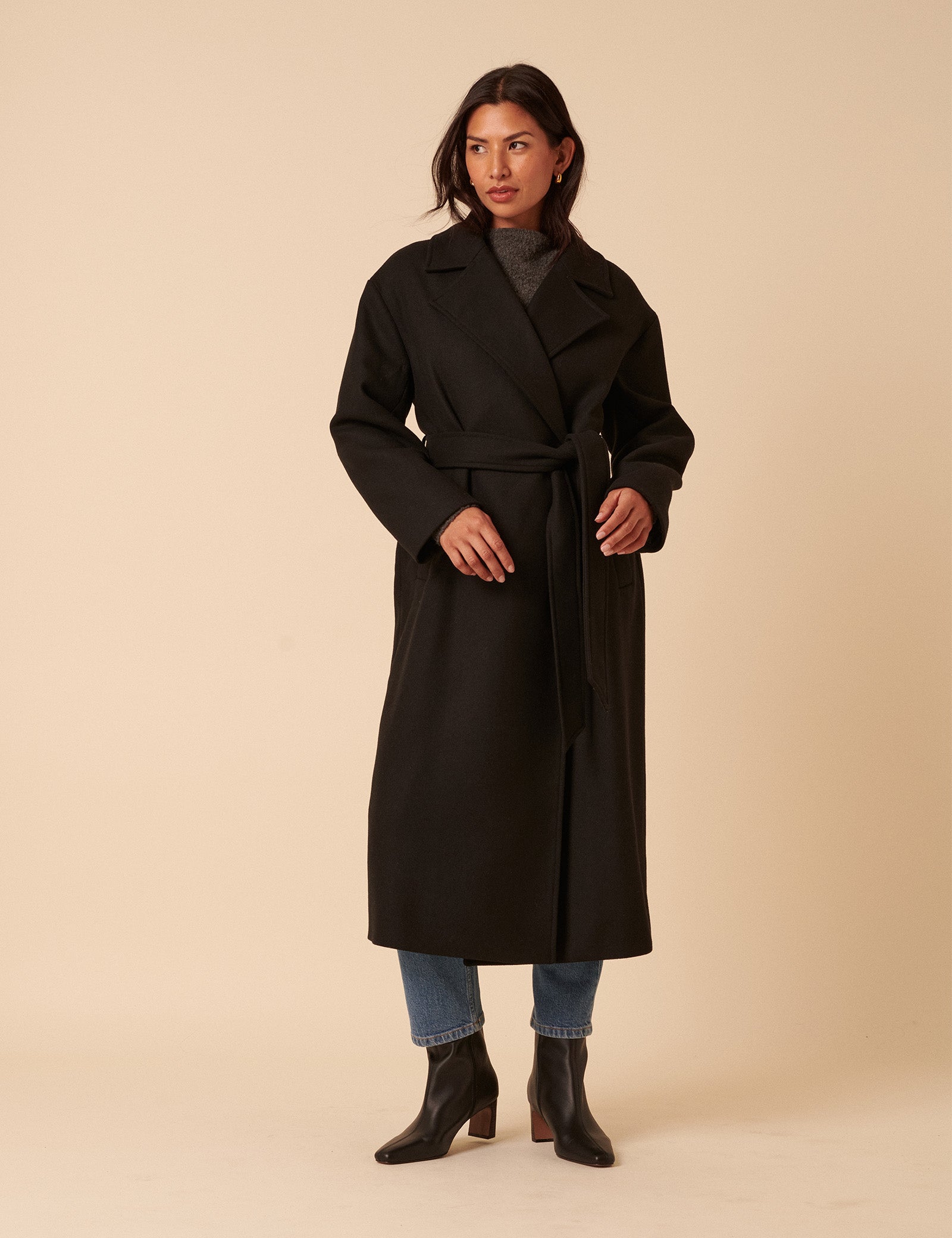 A woman wearing a long black wrap coat with a collar, long sleeves, side pockets, and a tie belt at the waist.