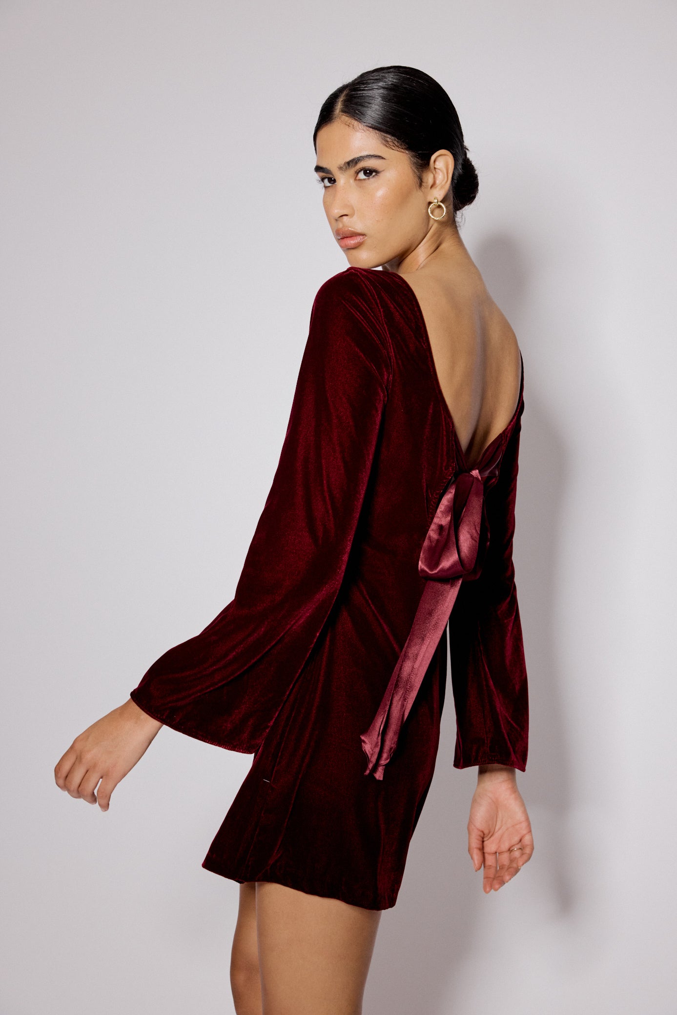 A woman wearing a burgundy satin mini dress with a boat neckline, long sleeves, and a bow at the back.