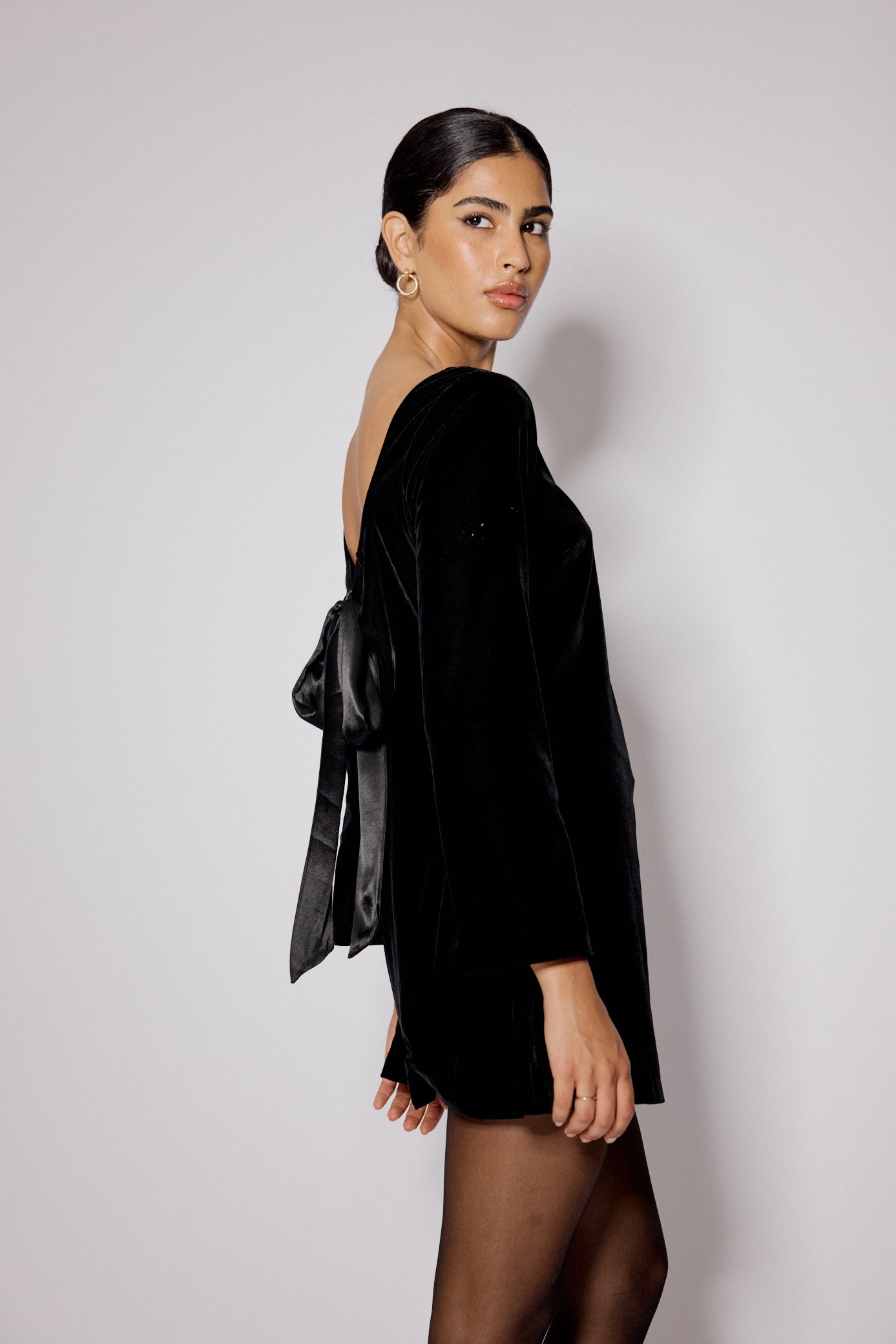 A woman wearing a black velvet mini dress with flare sleeves, boat neckline, and satin bow at the back.