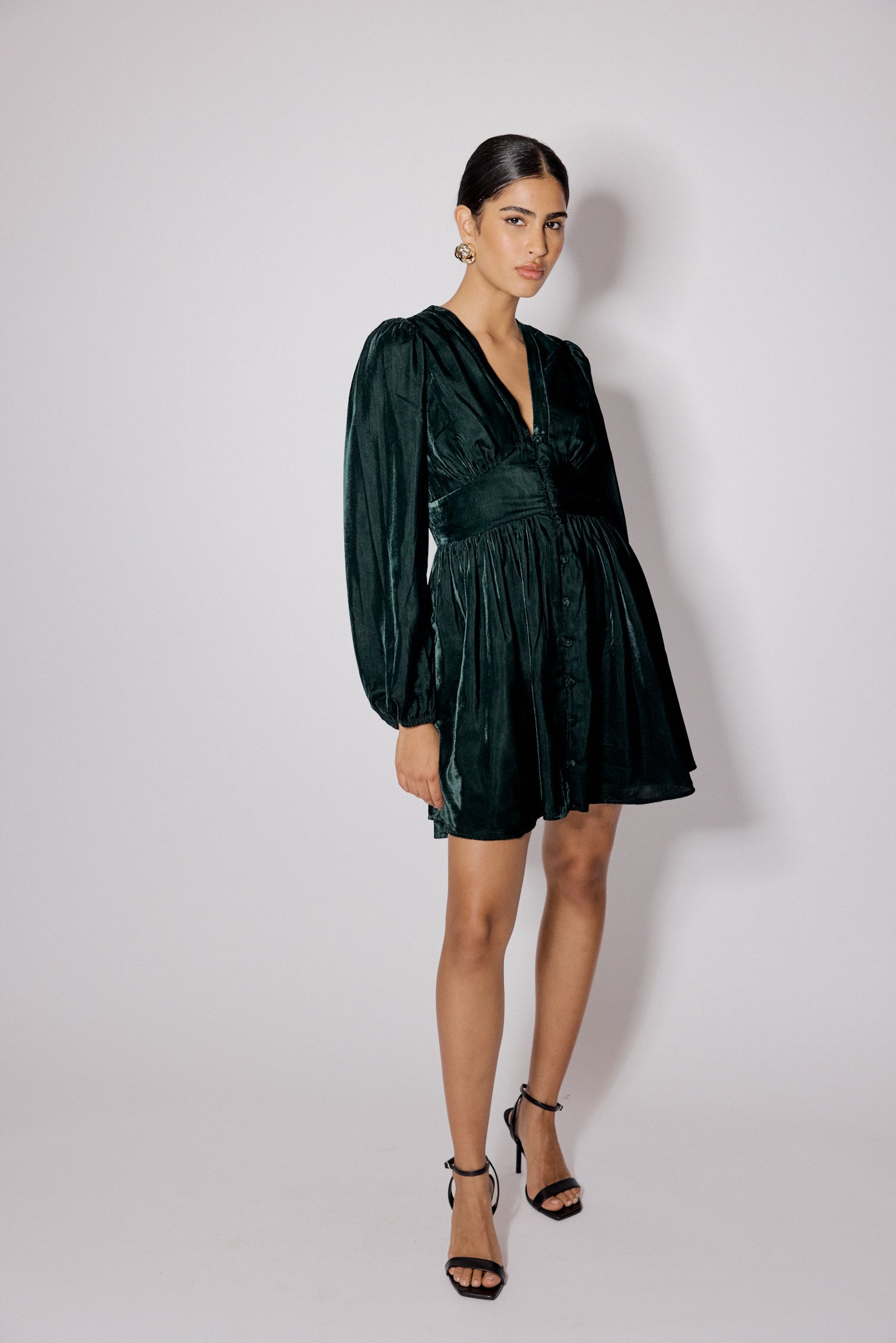 A woman wearing a green velvet mini dress with a V-neckline, long sleeves, and button details, standing against a plain background.
