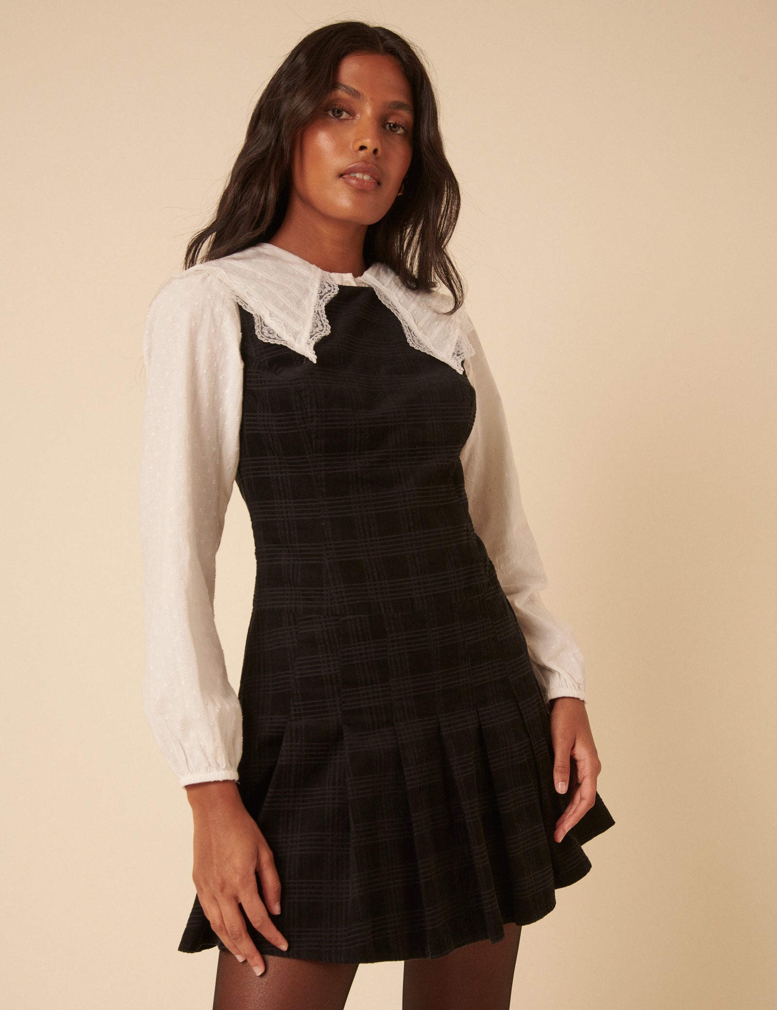 A woman wearing a black check cord Merida pinafore mini dress with a round neckline, sleeveless, drop waist, and pleated skirt.