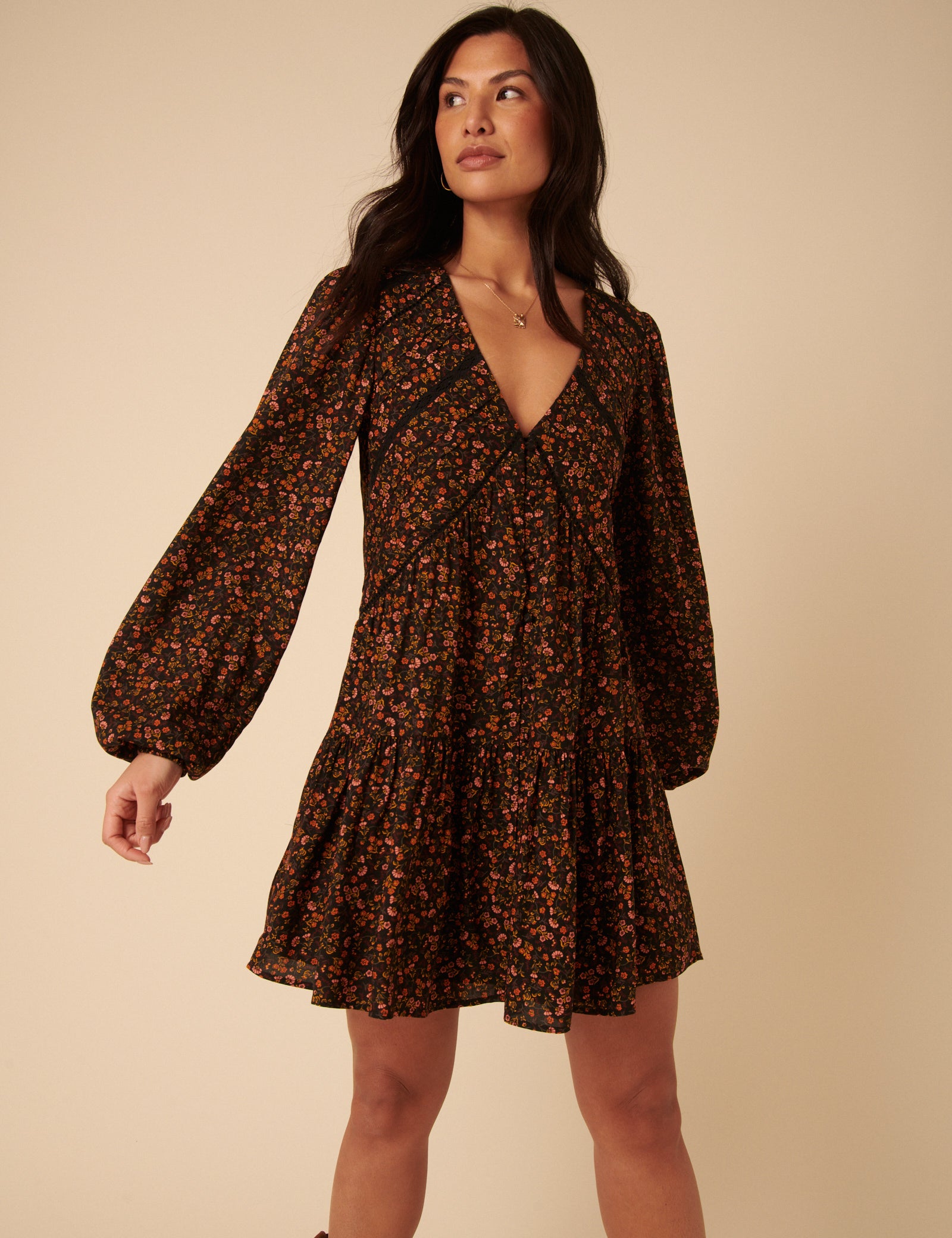 A woman wearing a black ditsy floral mini dress with long sleeves, V-neckline, and tiered hem.
