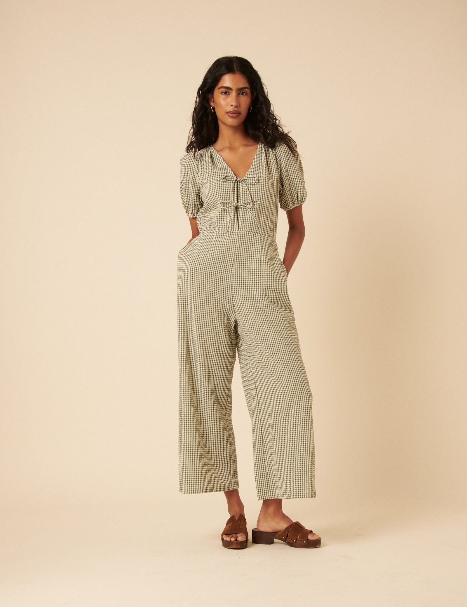 A woman wearing a green gingham jumpsuit with a V-neck, tie front, and wide-leg pants, standing against a plain background.