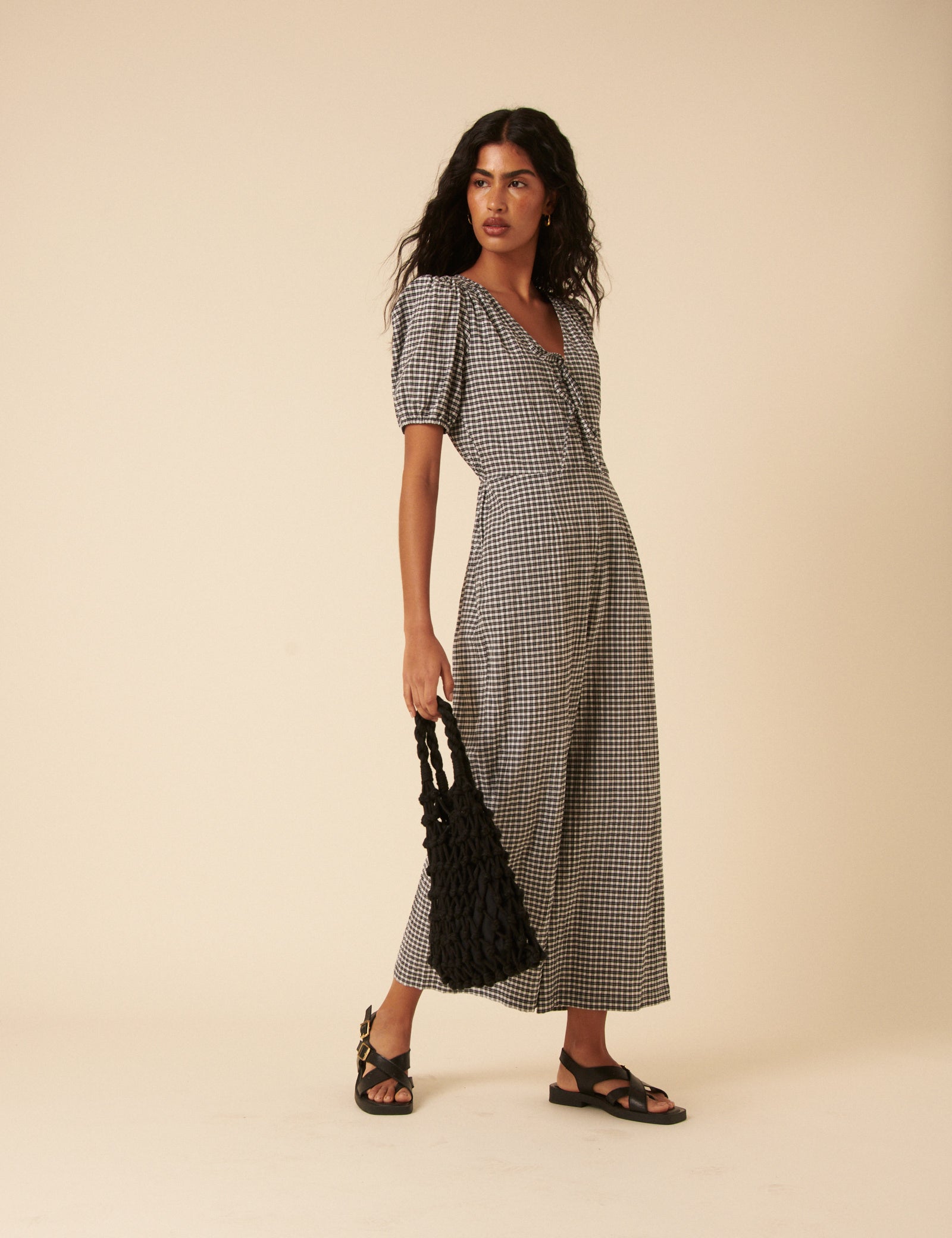 A woman wearing a black and white gingham jumpsuit with a V-neck, tie front, puffed sleeves, and wide-leg pants, holding a black woven bag and wearing black sandals.