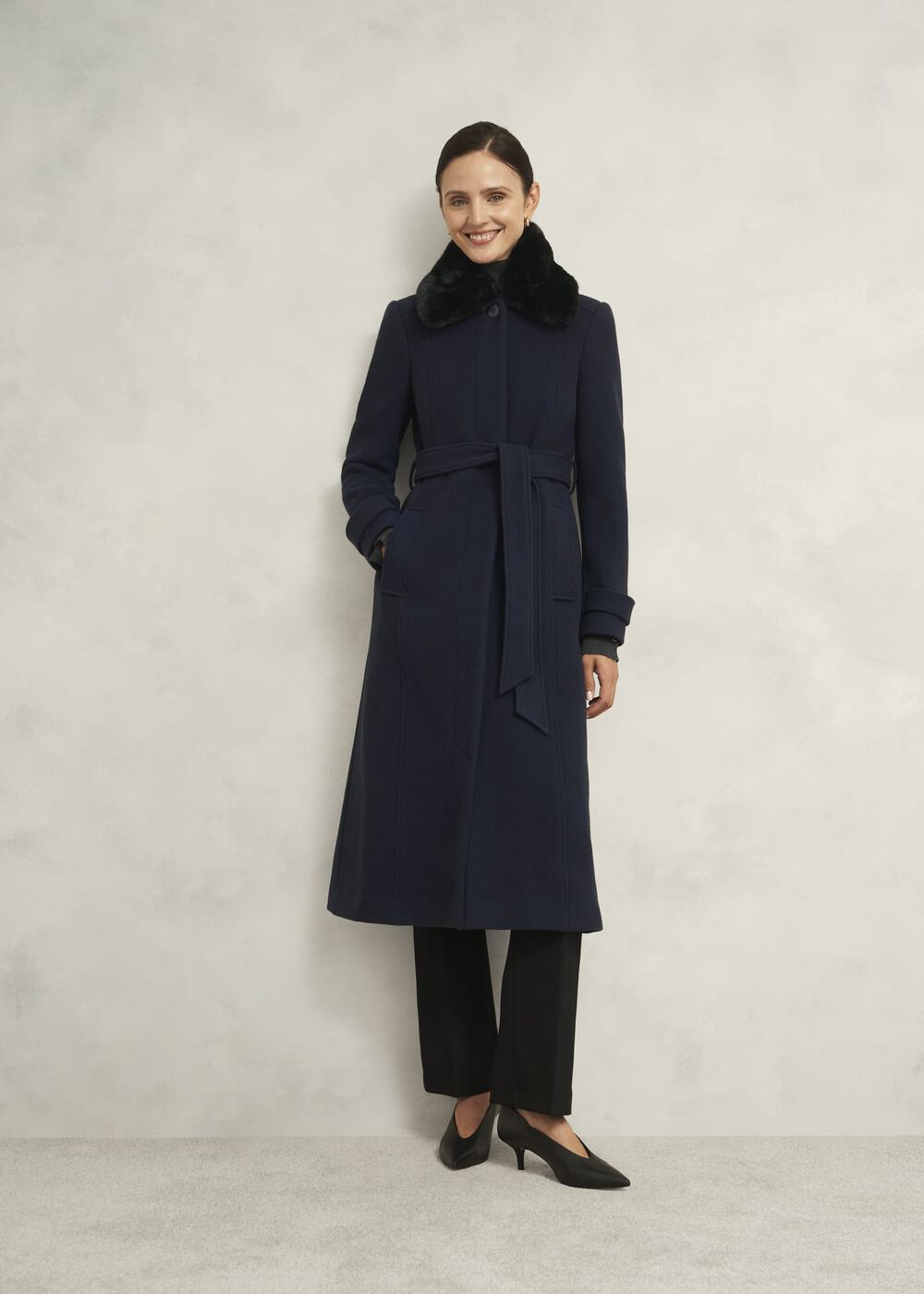A woman wearing a navy blue coat with a black faux fur collar, tie belt, and side pockets, standing against a plain background.