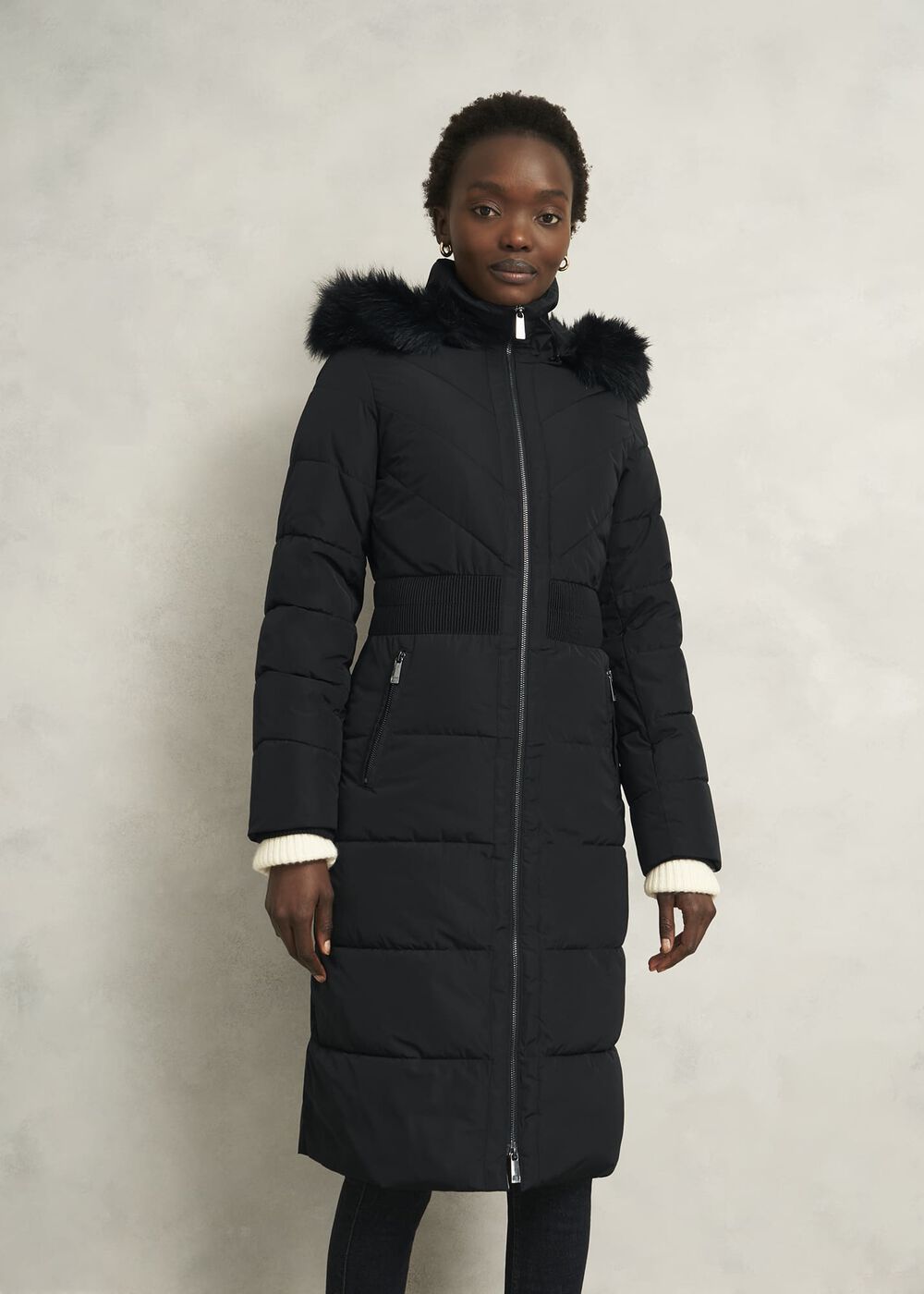 A woman wearing a long black puffer coat with a faux fur-trimmed hood, quilted pattern, and zippered pockets.