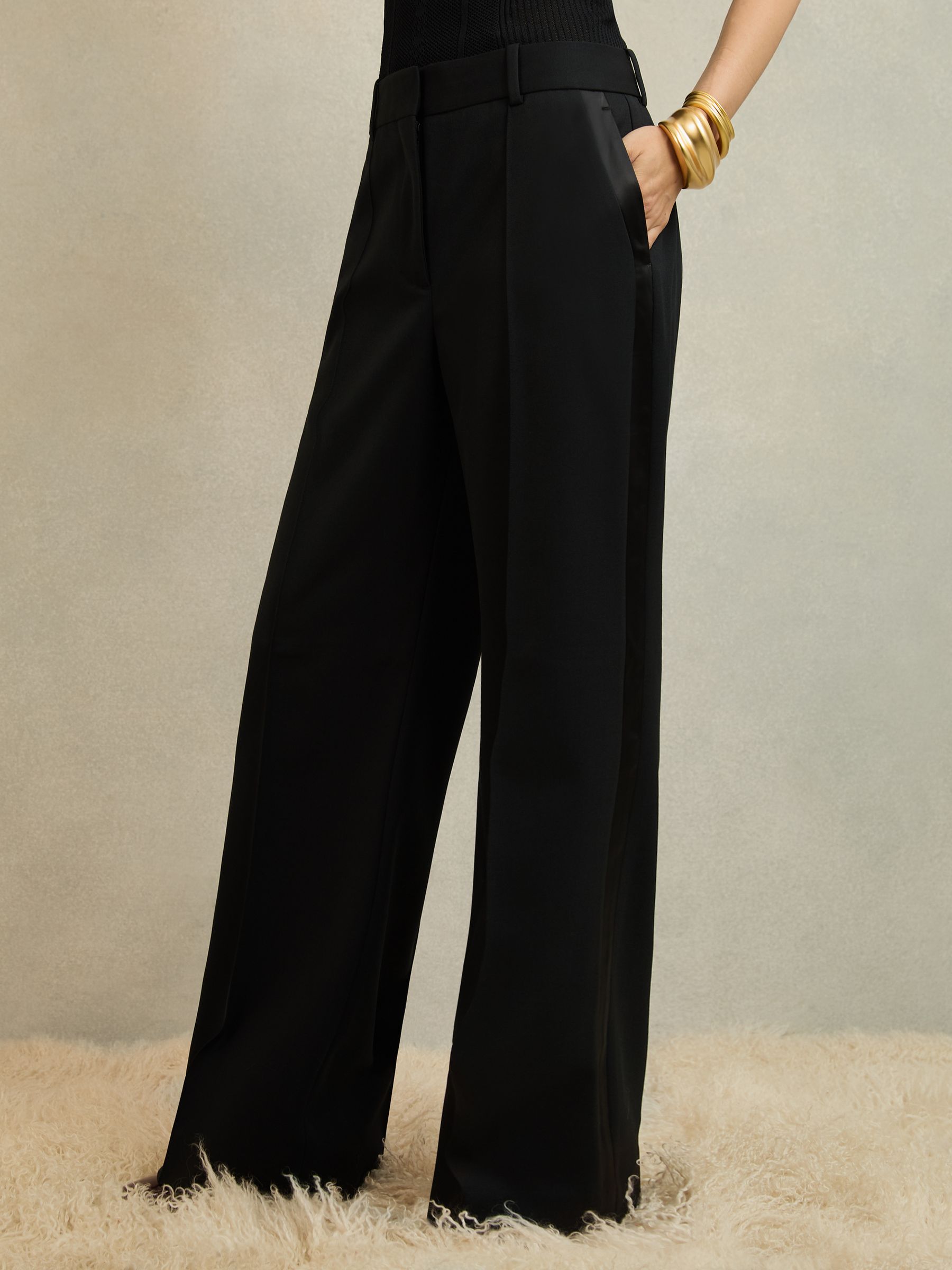 A pair of black wide-leg tuxedo trousers with satin stripe along the side seam, featuring sharp pintucks and angled side pockets.