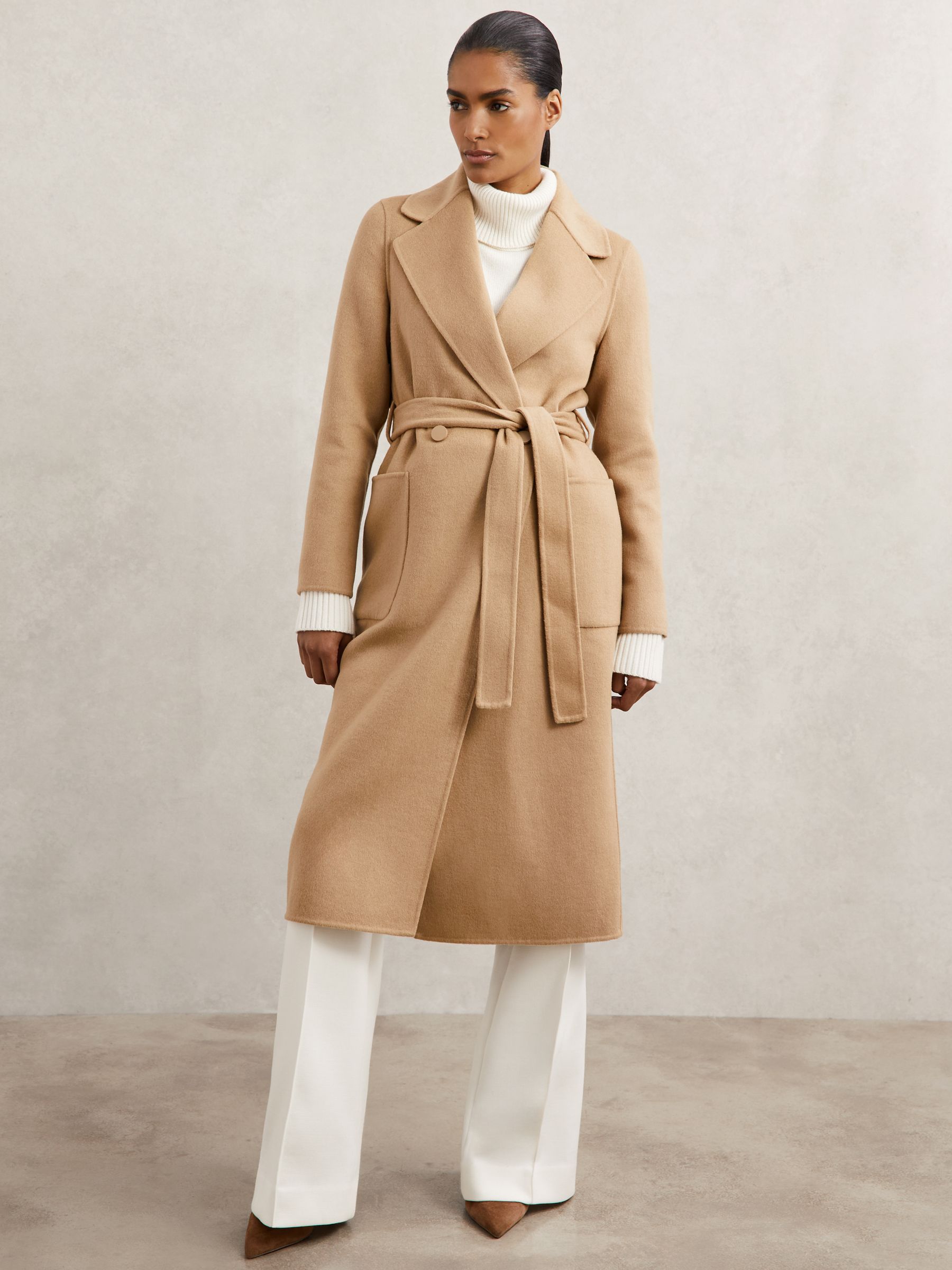 A woman wearing a camel-colored, double-breasted wool-blend coat with a self-tie belt and blind seams, standing against a plain background.