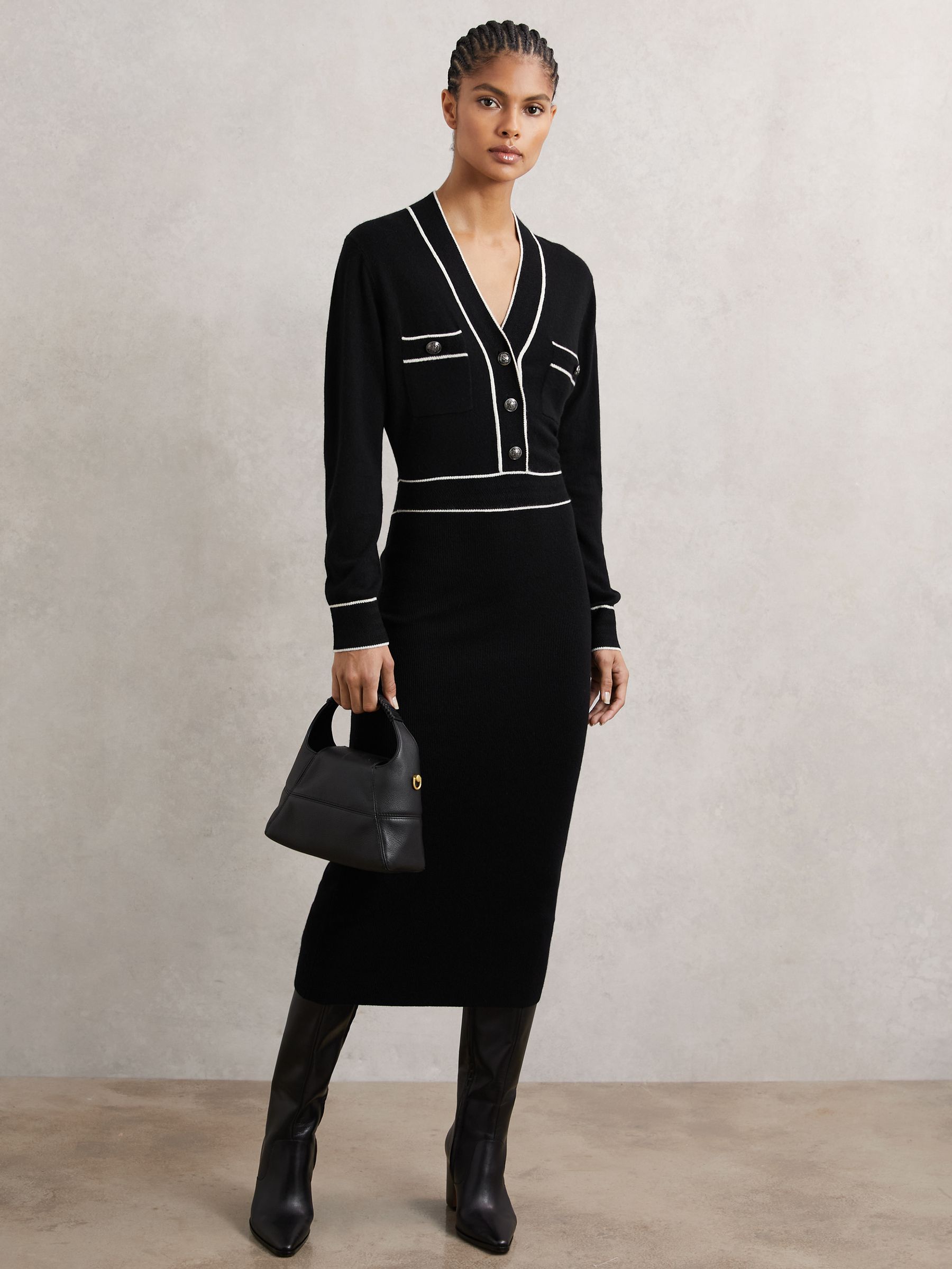 A woman wearing a black wool-blend dress with white tipping details, crest-embossed buttons, and a ribbed midi skirt, holding a black handbag and wearing black knee-high boots.