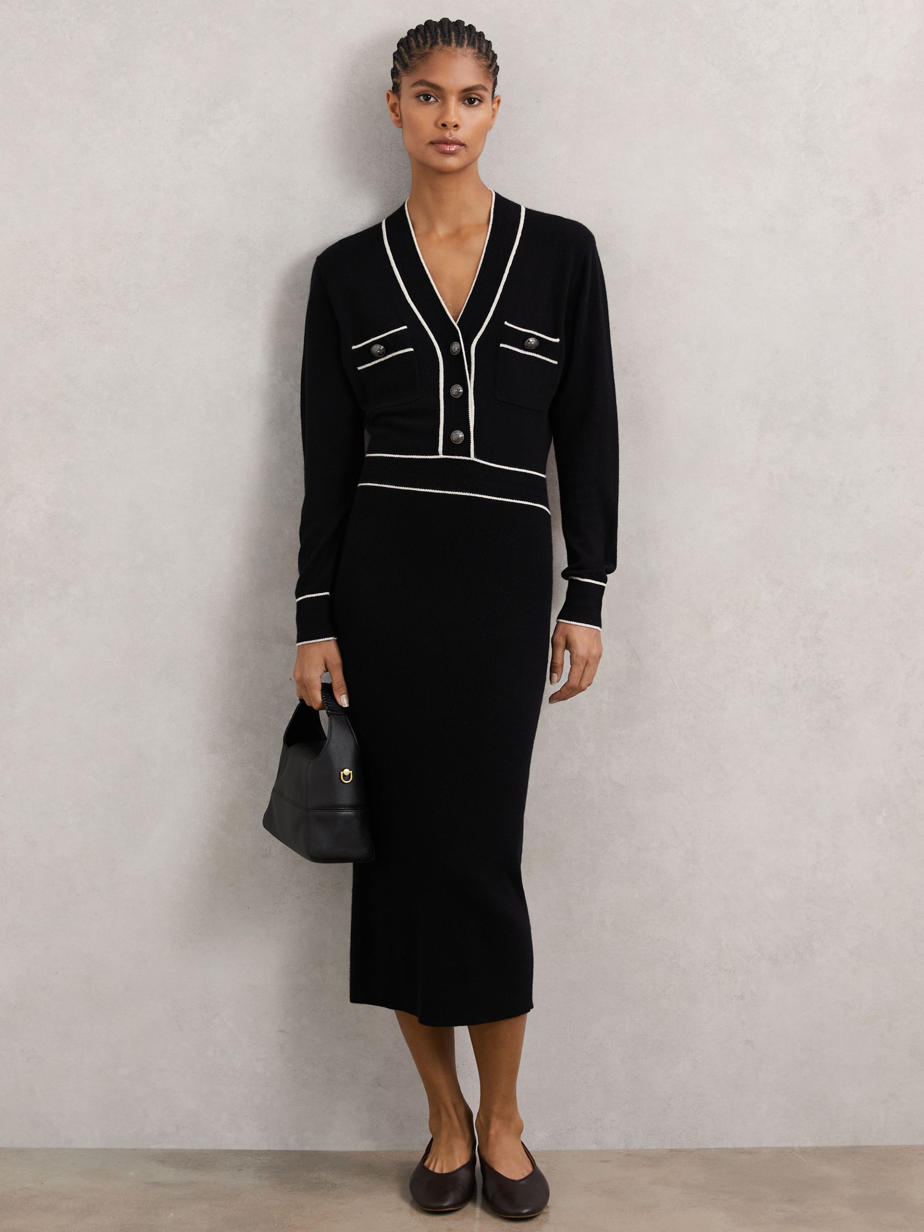 A woman wearing a black knitted dress with ivory tipped accents, crest-embossed buttons, and a ribbed midi skirt.
