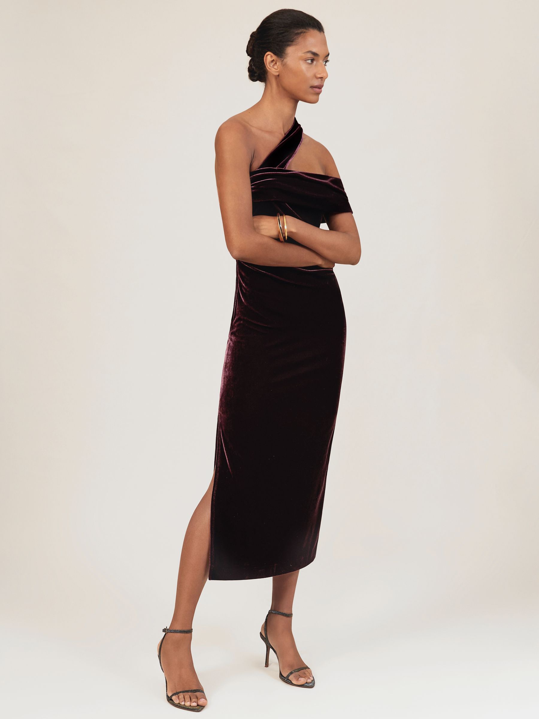 A woman wearing a burgundy velvet one-shoulder midi dress with ruching and a side slit.