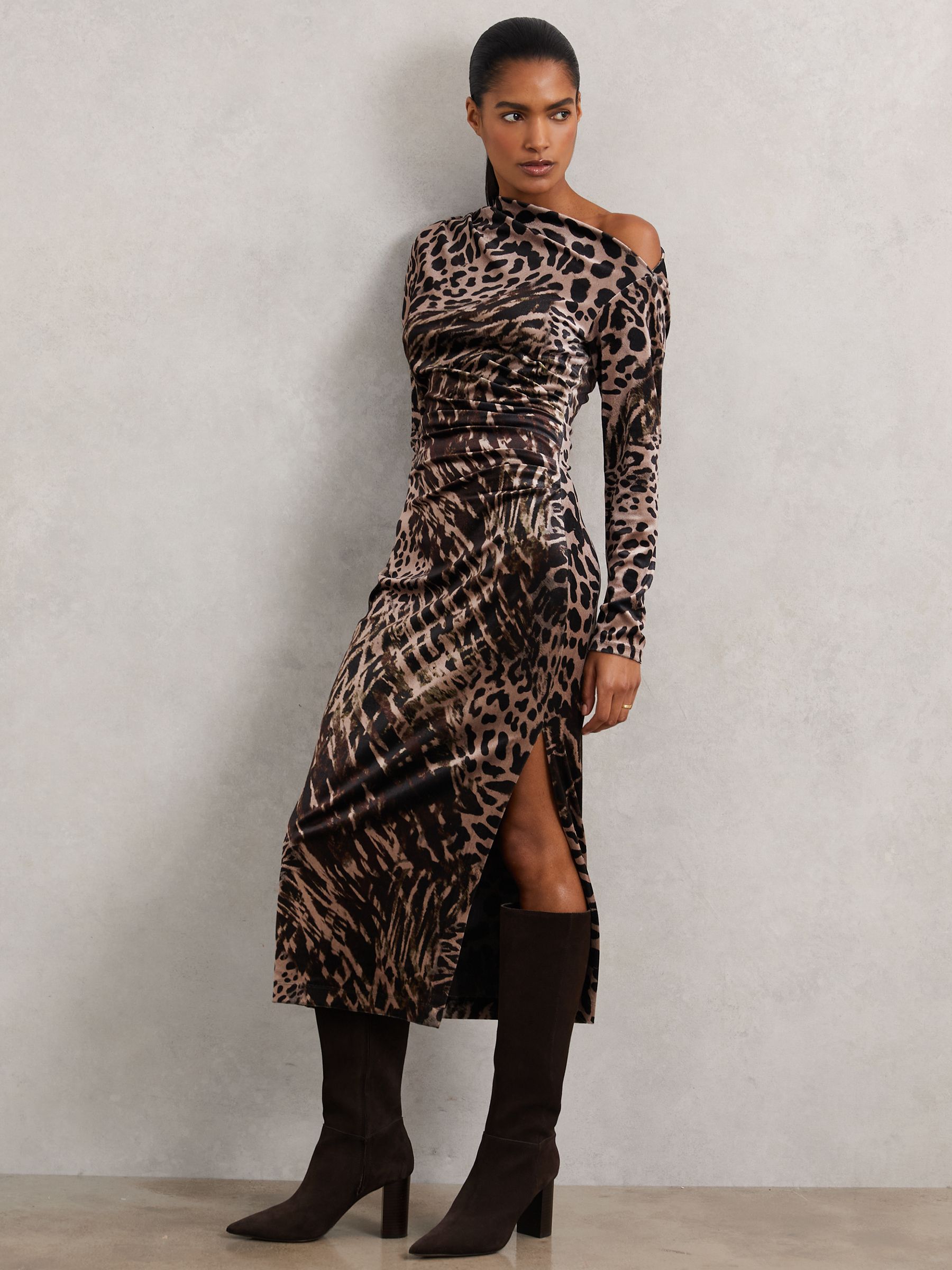 A woman wearing a leopard-print midi dress with an asymmetric off-shoulder neckline, ruched waist, and high side slit, paired with knee-high black boots.