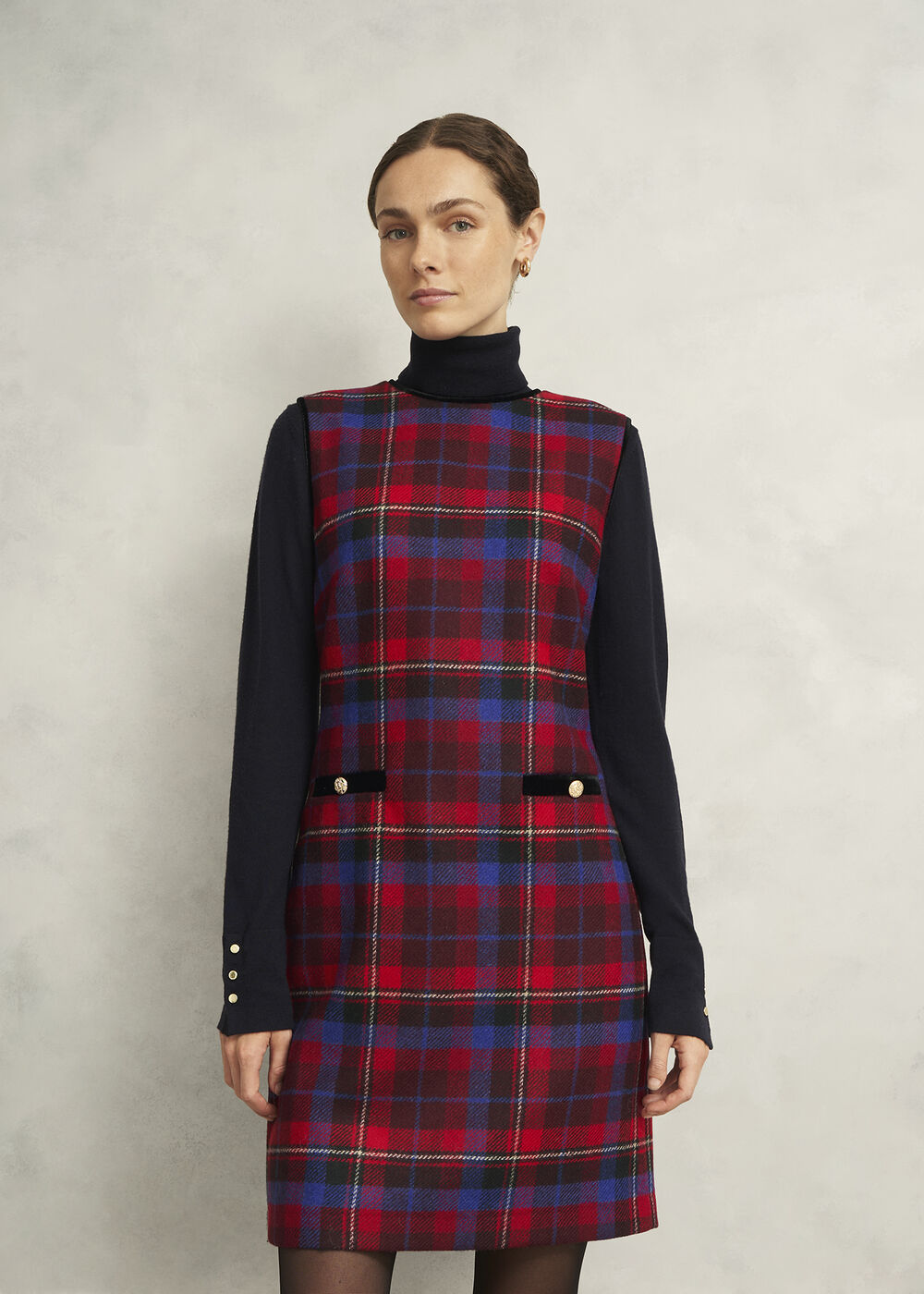 A woman wearing a red, blue, and black heritage check wool dress with velvet trims and pocket details, styled as a short shift dress.