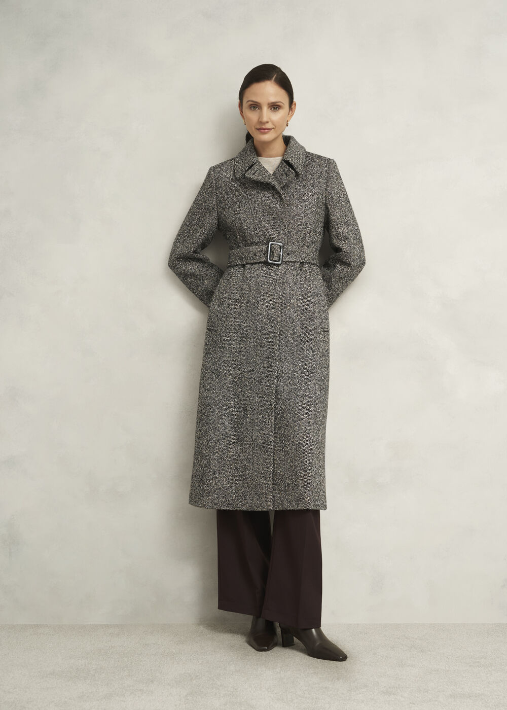 A woman wearing a salt-and-pepper wool blend coat with a tailored fit, belt, side pockets, and asymmetric seams.