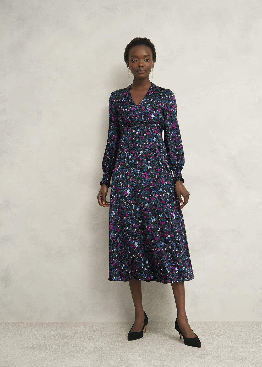 A woman wearing a long, floral print dress with semi-sheer long sleeves, a fitted waist, and a flared skirt, paired with black high heels.