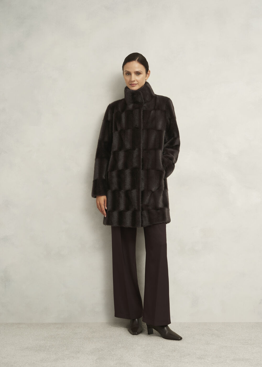 A woman wearing a dark brown faux fur coat with a longline, boxy fit, standing against a plain background.