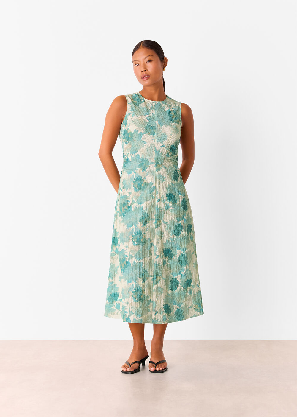 A woman wearing a sleeveless, textured midi dress with a floral pattern in shades of blue and cream, round neckline, and ruched sides.