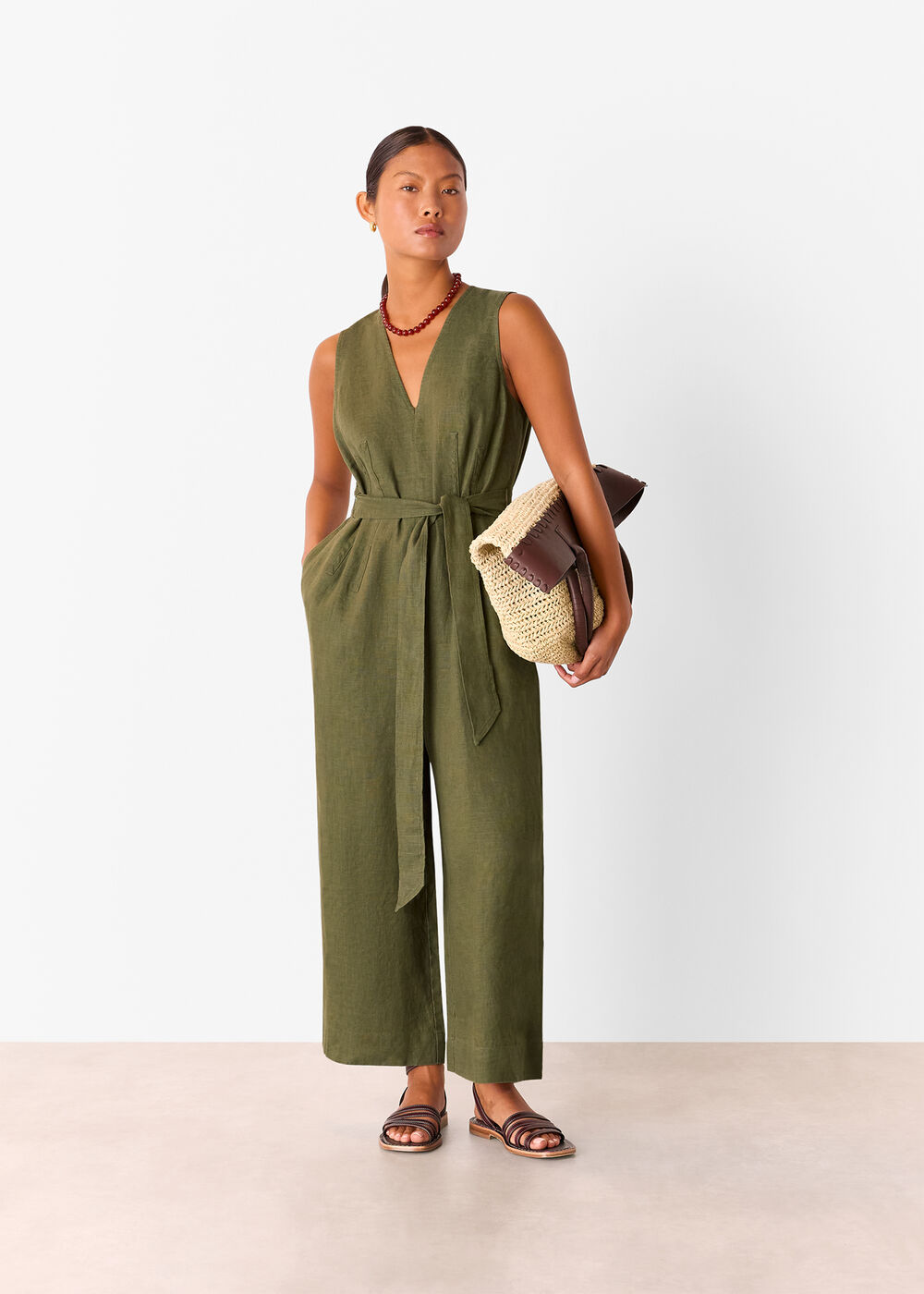 A woman wearing a khaki linen jumpsuit with a V-neckline, belt at the waist, and ankle-length hem, holding a woven bag and wearing sandals.