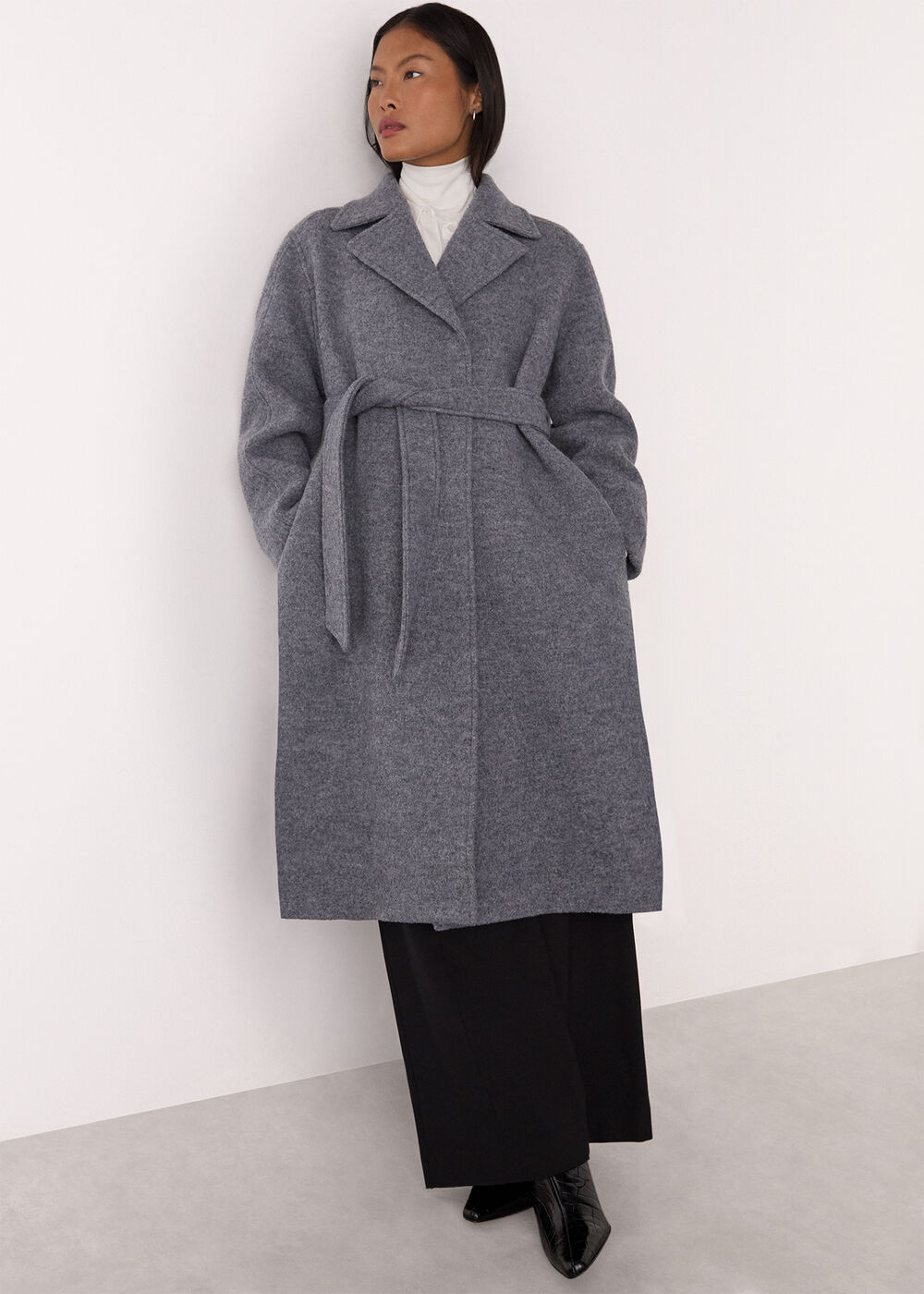 A woman wearing a long grey wool coat with a belt, collared neckline, and two pockets, standing against a plain white background.