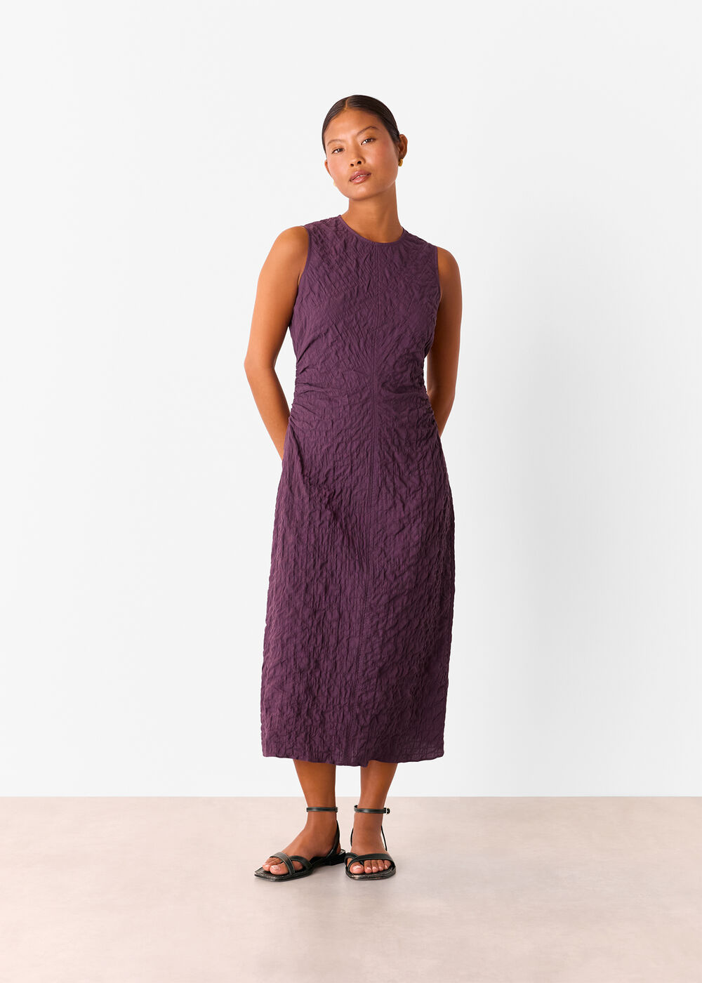 A woman wearing a sleeveless plum-colored textured midi dress with ruched detailing, round neckline, and black sandals.