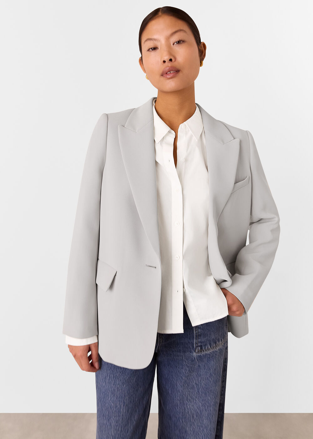 A woman wearing a light beige blazer with peak lapels, a button closure, and a relaxed, boxy fit, paired with a white shirt and blue jeans.