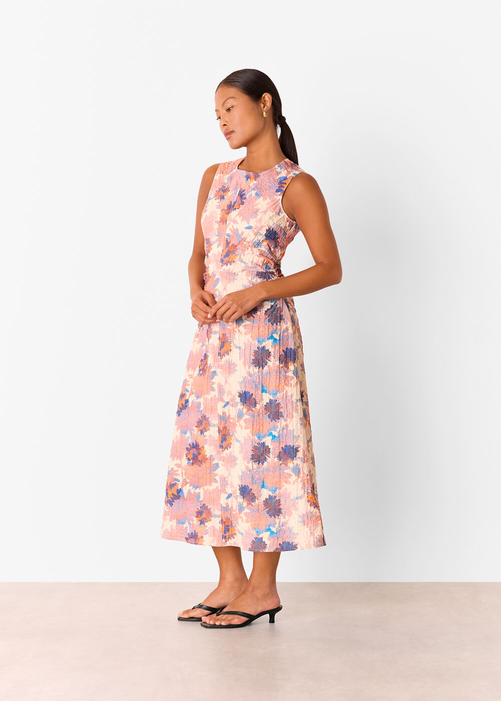 A woman wearing a pink floral textured midi dress with a sleeveless design, round neckline, and ruched sides, standing against a plain background.