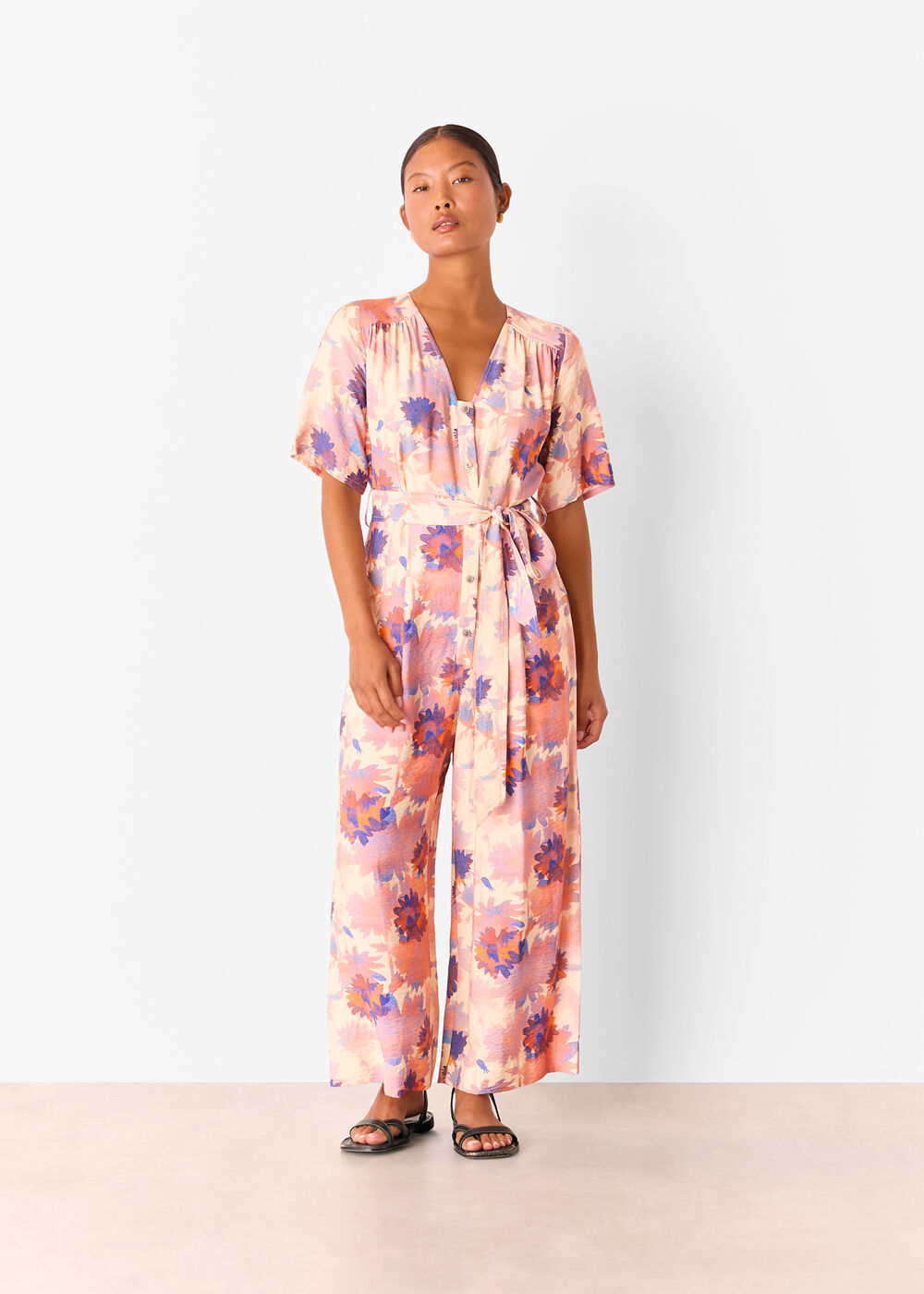 A woman wearing a pink petite floral jumpsuit with short sleeves, a V-neckline, and a belt, standing against a plain background.