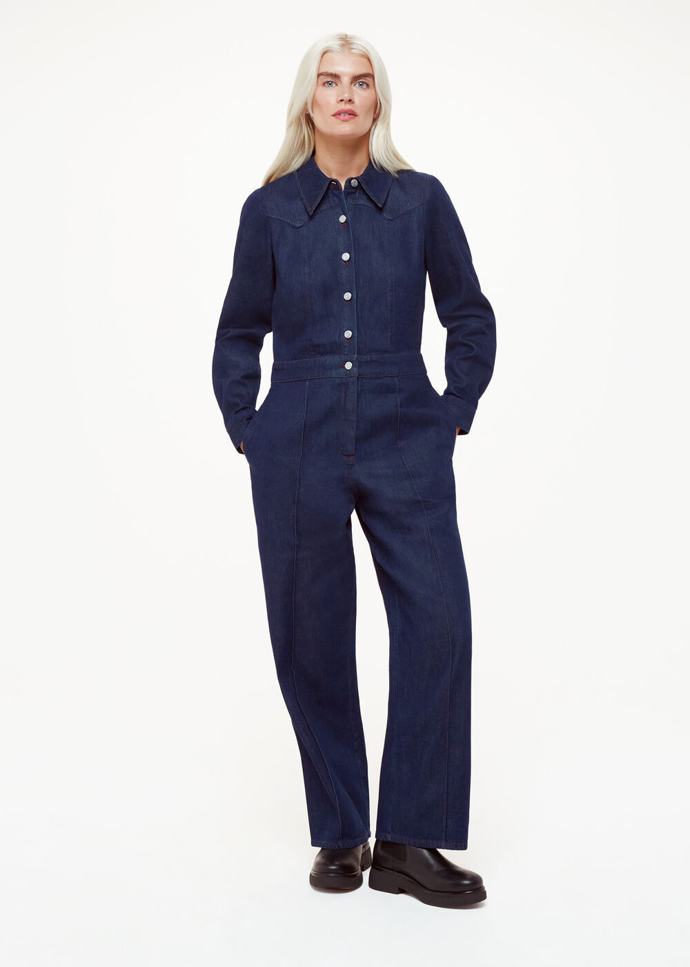 A woman wearing a dark blue denim jumpsuit with a collar, long sleeves, and wide-leg pants, standing with her hands in her pockets.
