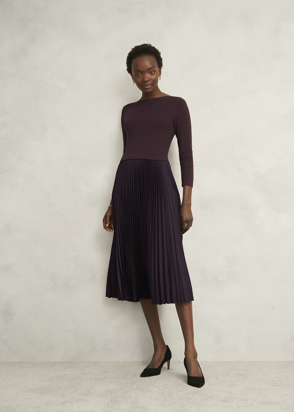 A woman wearing a deep purple knitted top with long slim sleeves and a boat neckline, paired with a matching pleated satin midi skirt.