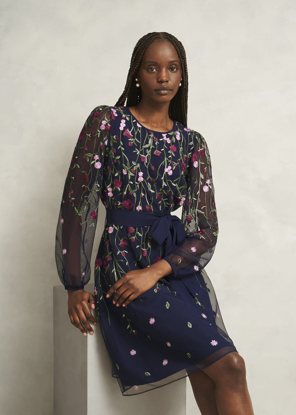 A woman wearing a navy embroidered mini dress with sheer long sleeves, floral embroidery, and a tie waist.