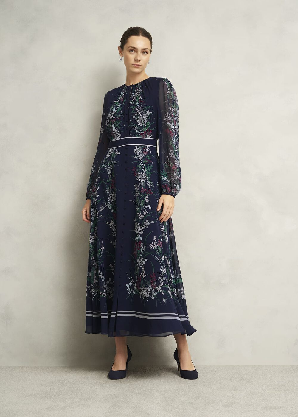 A woman wearing a navy blue floral silk dress with semi-sheer sleeves, gathered neckline, and button details, paired with black heels.