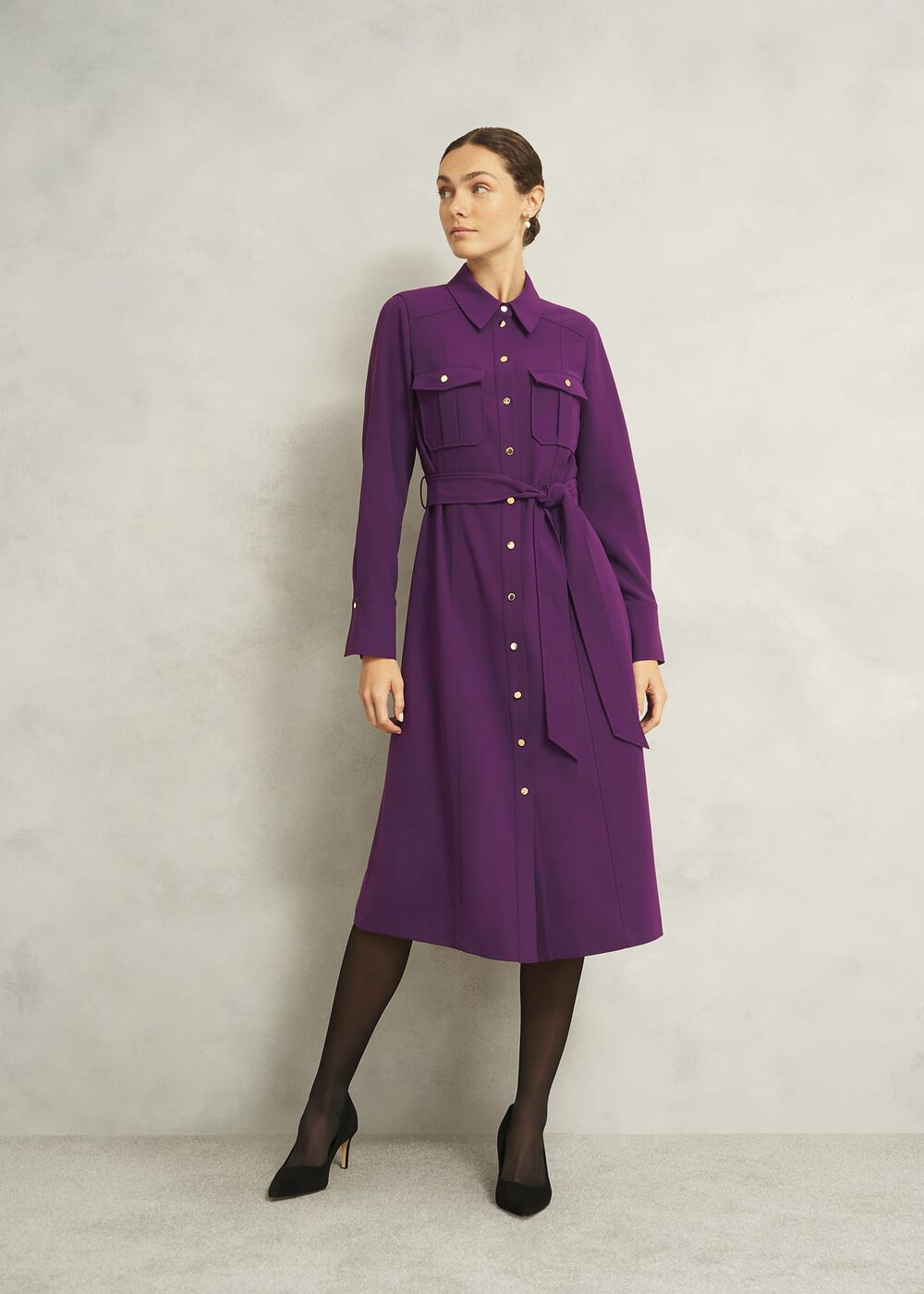 A woman wearing a vibrant purple shirt dress with long sleeves, button-down front, tie waist, and side pockets, paired with black heels and sheer black tights.