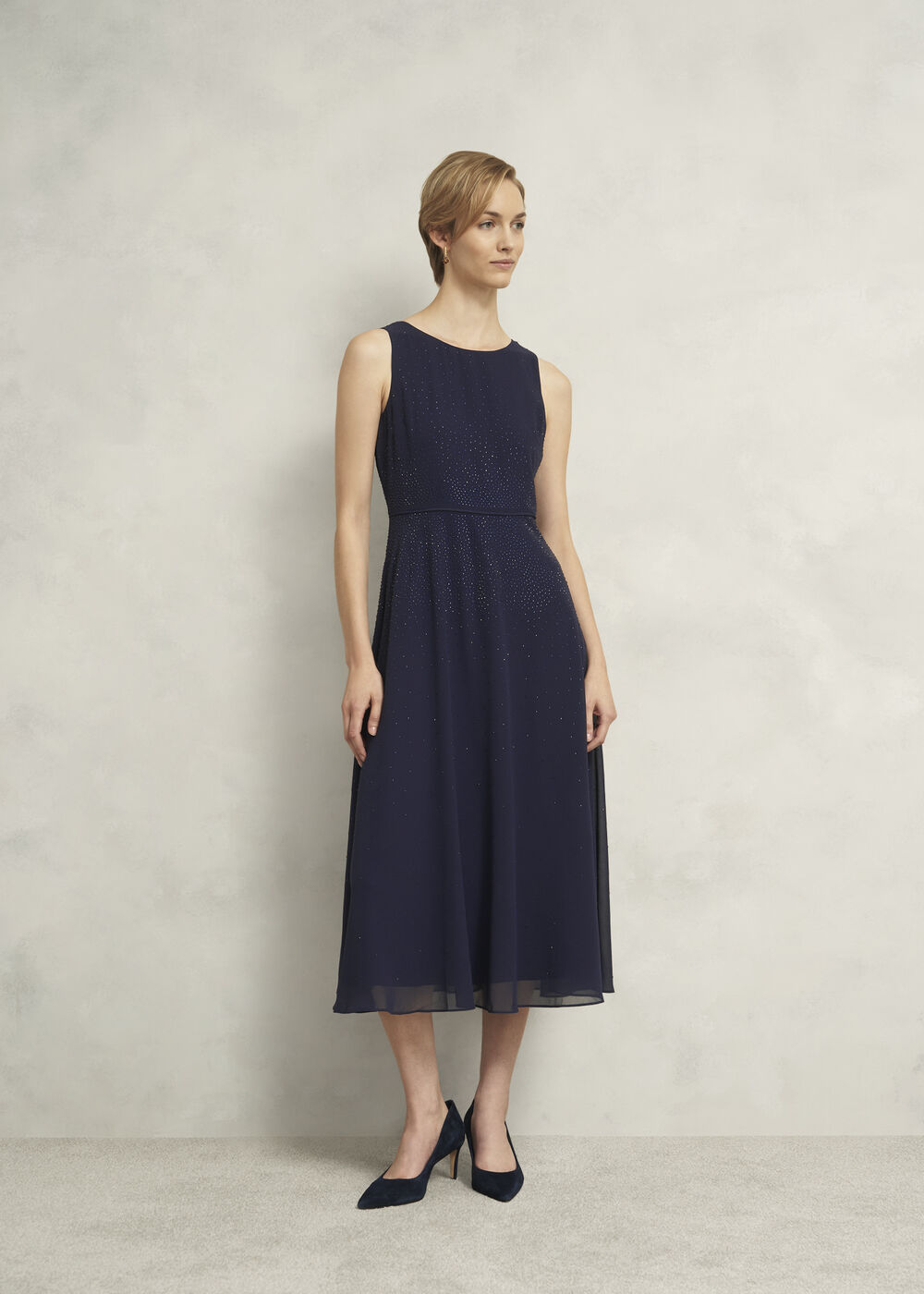 A woman wearing a navy blue sleeveless chiffon dress with tonal gems and a keyhole back, paired with navy heels.