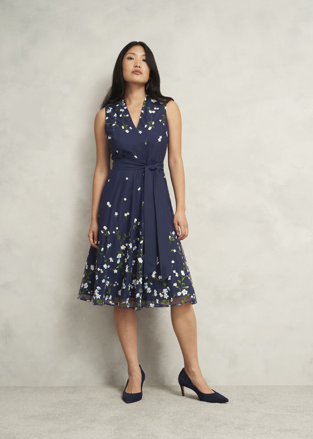 A woman wearing a navy blue floral embroidered sleeveless dress with a waist tie, V neckline, and a flared midi length.