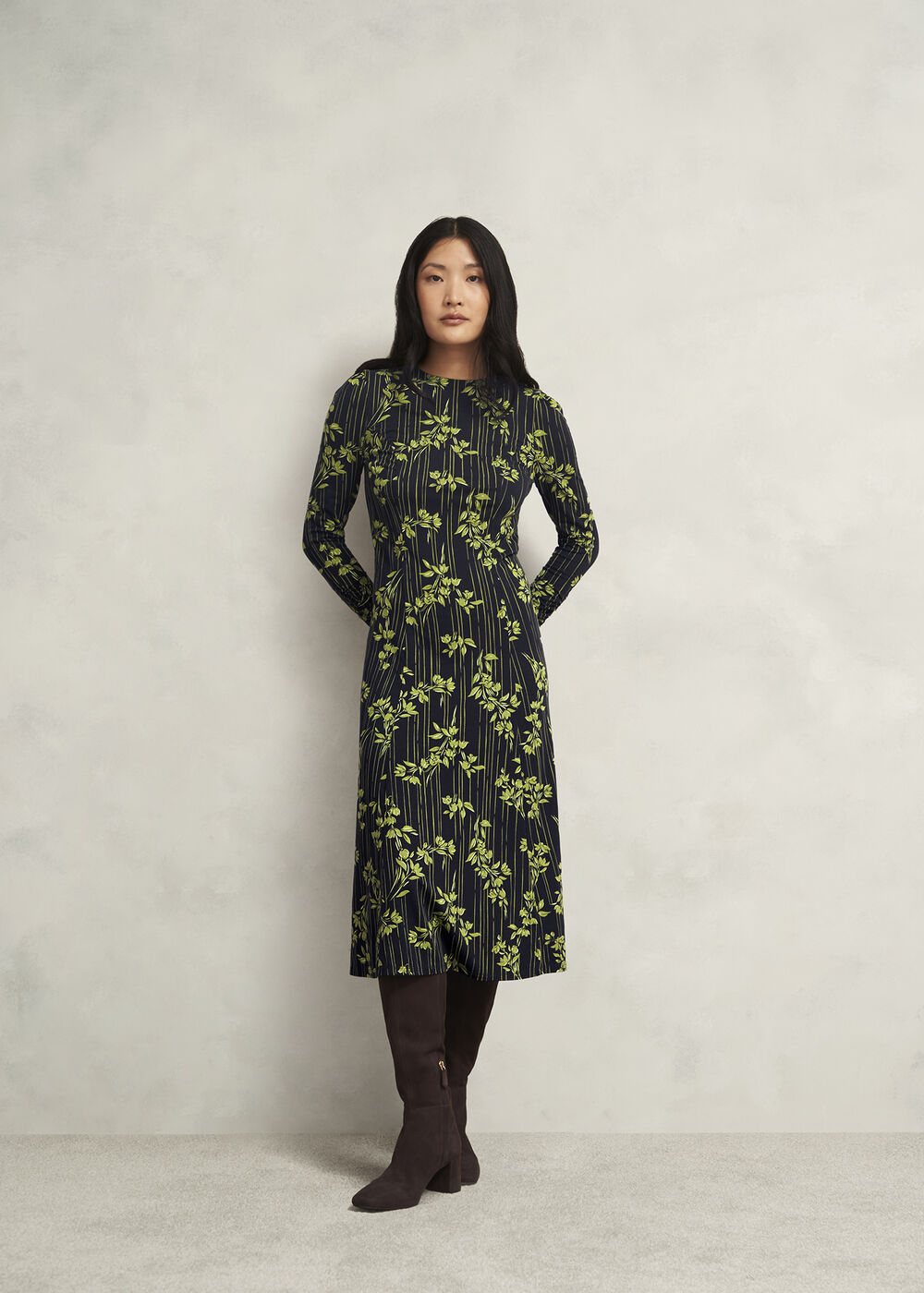 A woman wearing a black midi dress with green botanical print, long sleeves, and a back zip, standing against a plain background.