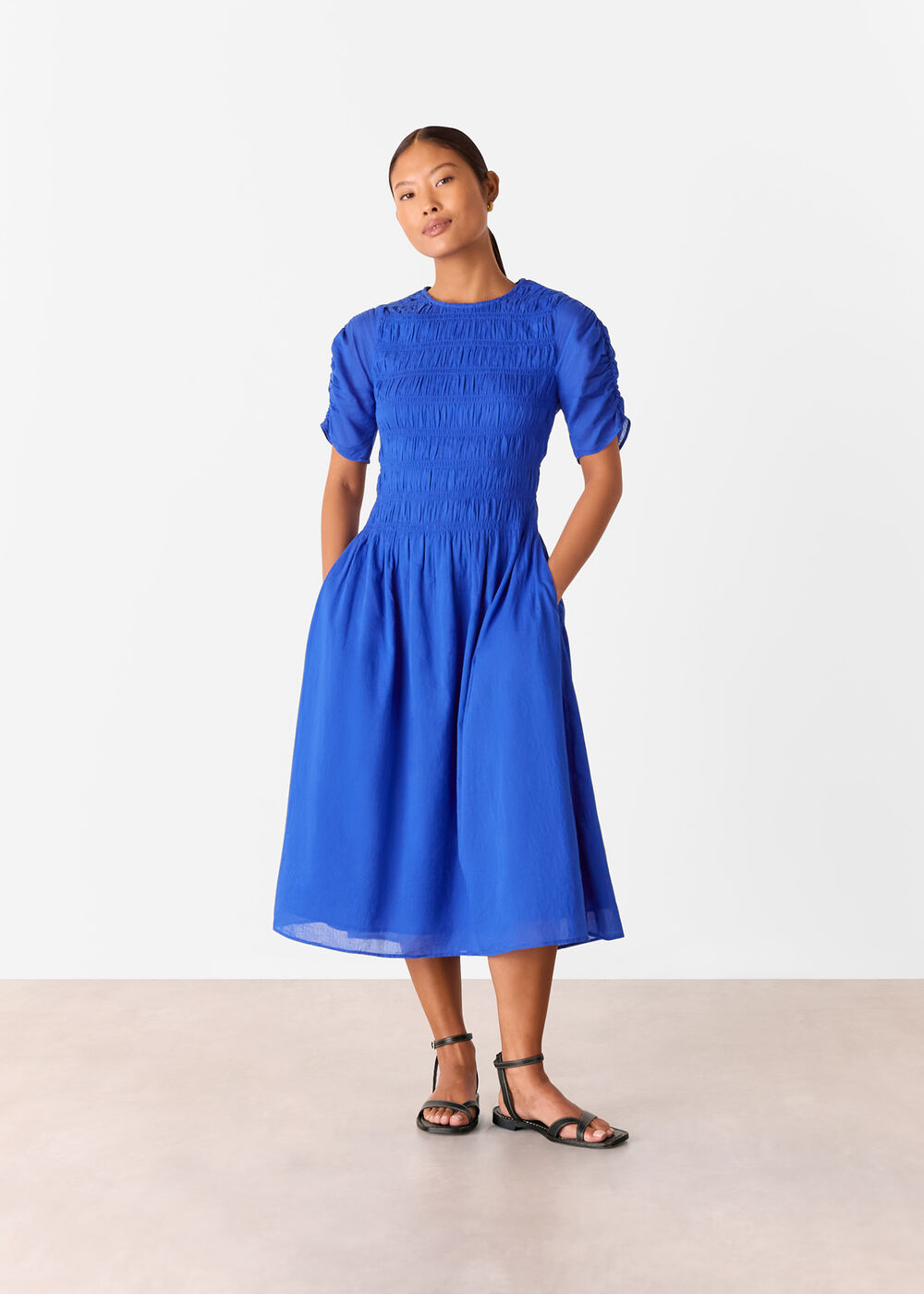 A woman wearing a bright blue smocked dress with short puff sleeves and a flared midi skirt, standing against a plain white background.