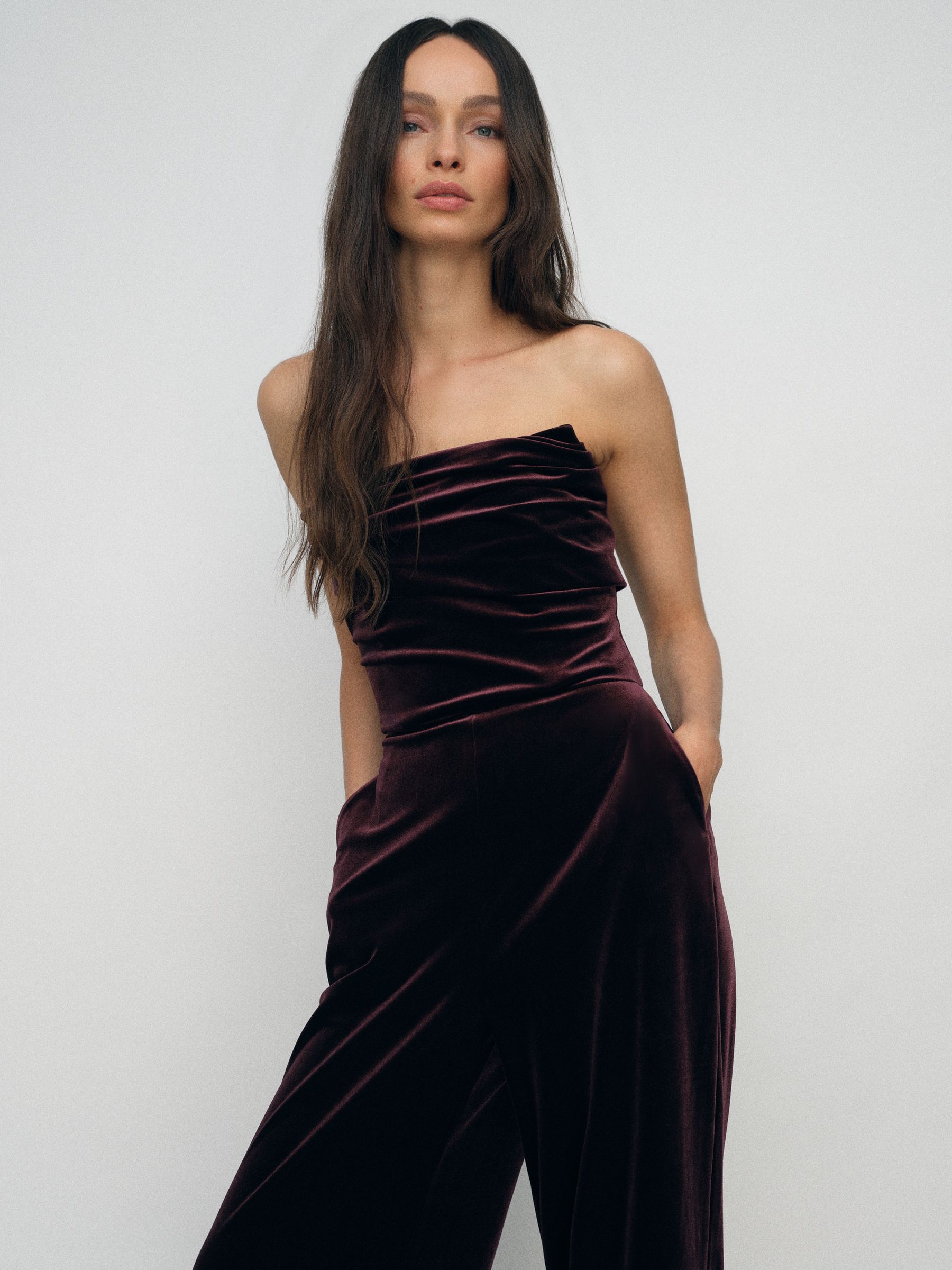 A woman wearing a strapless burgundy velvet jumpsuit with a draped neckline and wide-leg trousers.