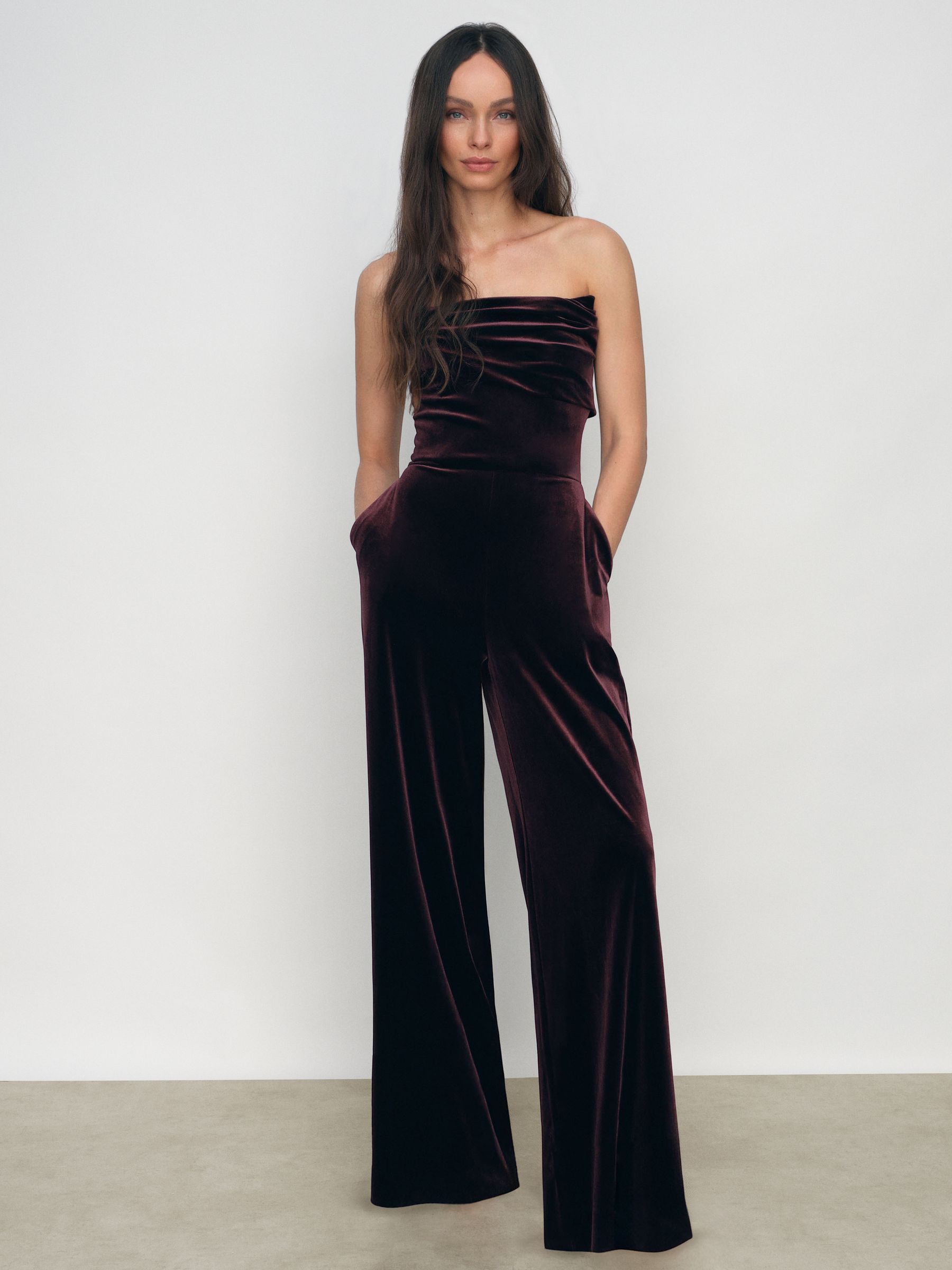A woman wearing a burgundy velvet strapless jumpsuit with a draped neckline and wide-leg trousers.