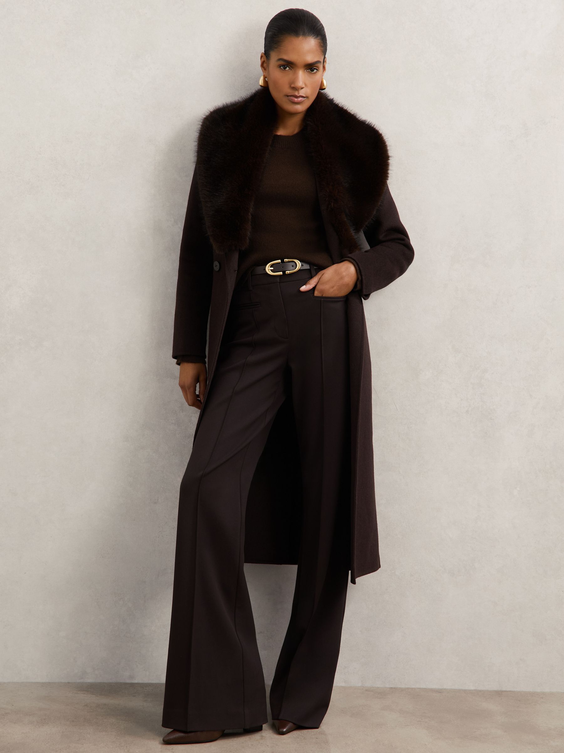 A woman wearing a chocolate brown wool-blend coat with a faux-fur collar, double-breasted style, and a self-tie belt, standing against a plain background.
