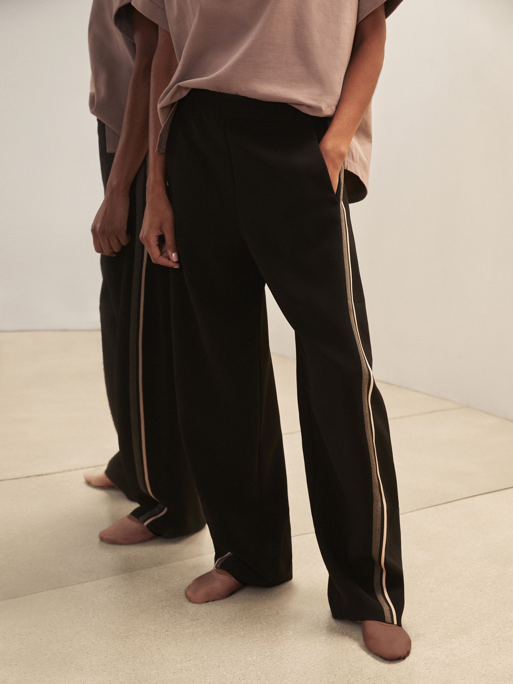 A pair of black wide-leg trousers with beige side stripes, elastic waistband, and a fluid woven fabric, worn by a person with their hand in the pocket.