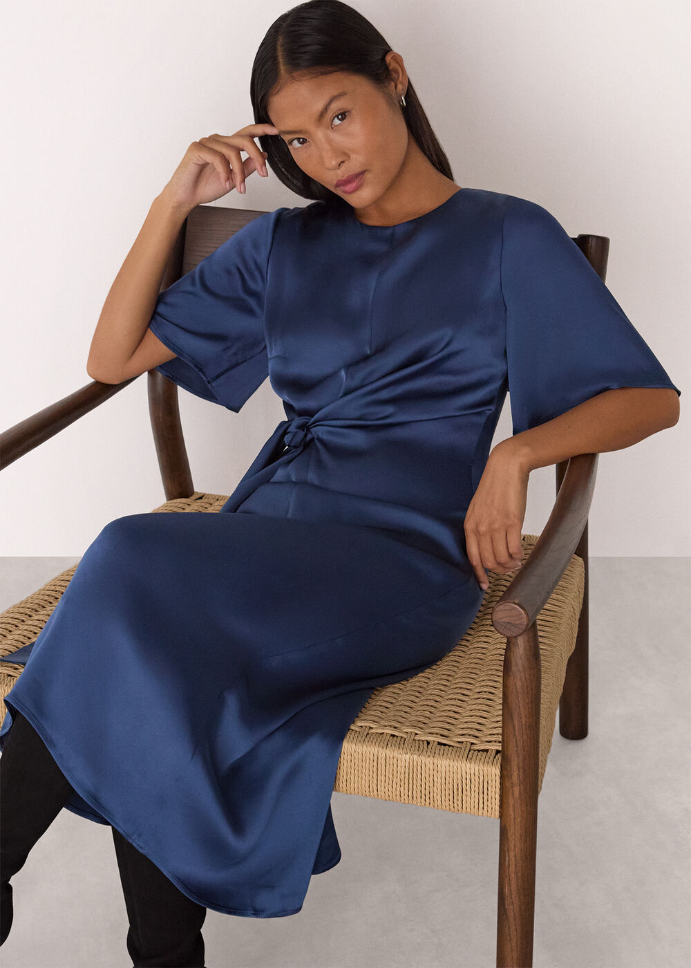 A woman wearing a blue satin midi dress with short sleeves and a twist at the waist, sitting on a wooden chair.