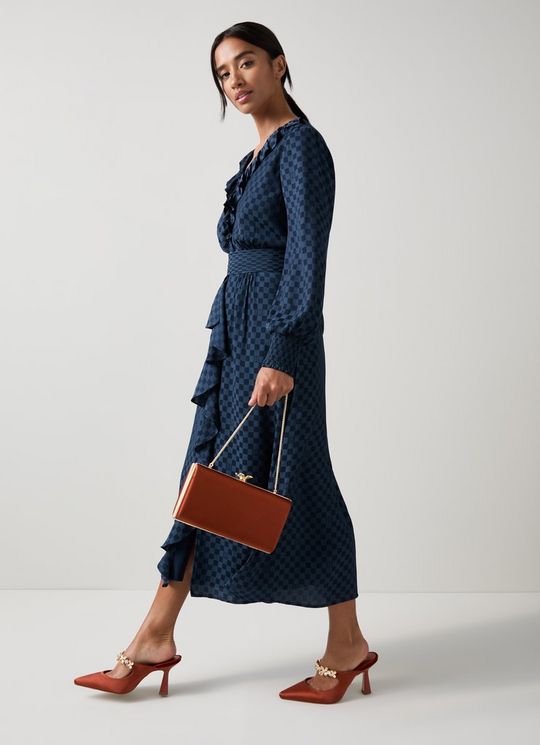 A woman wearing a navy blue midi dress with a split neckline, long blouson sleeves, and waterfall frill details, paired with brown heels and a brown handbag.