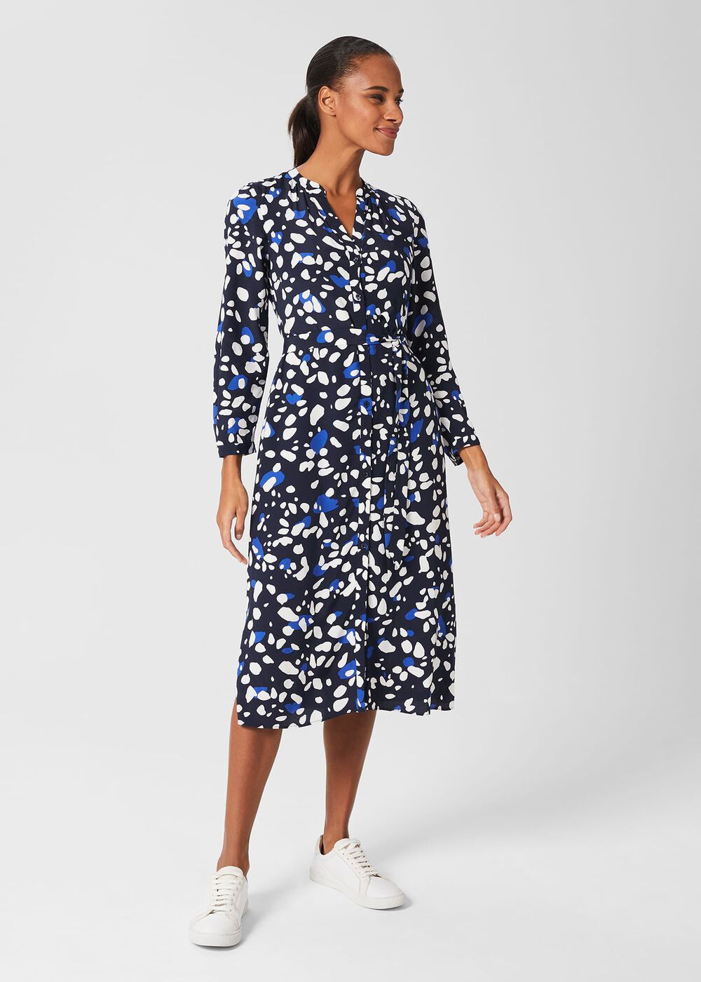 A woman wearing a navy blue midi dress with a white and blue abstract print, long sleeves, a tie waist, and side pockets, styled with white sneakers.
