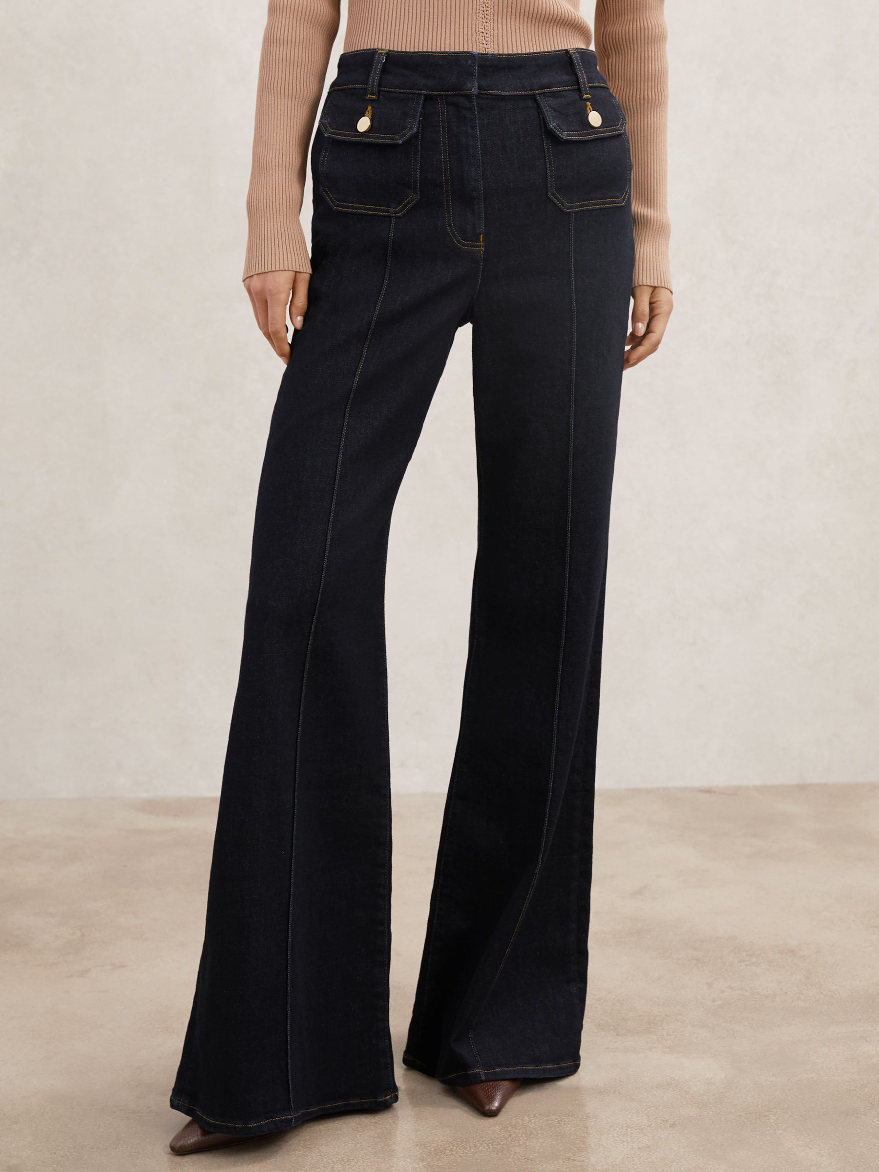 A pair of dark blue petite flared jeans with pocket details, wide hem, and button-flap pockets at the waist.